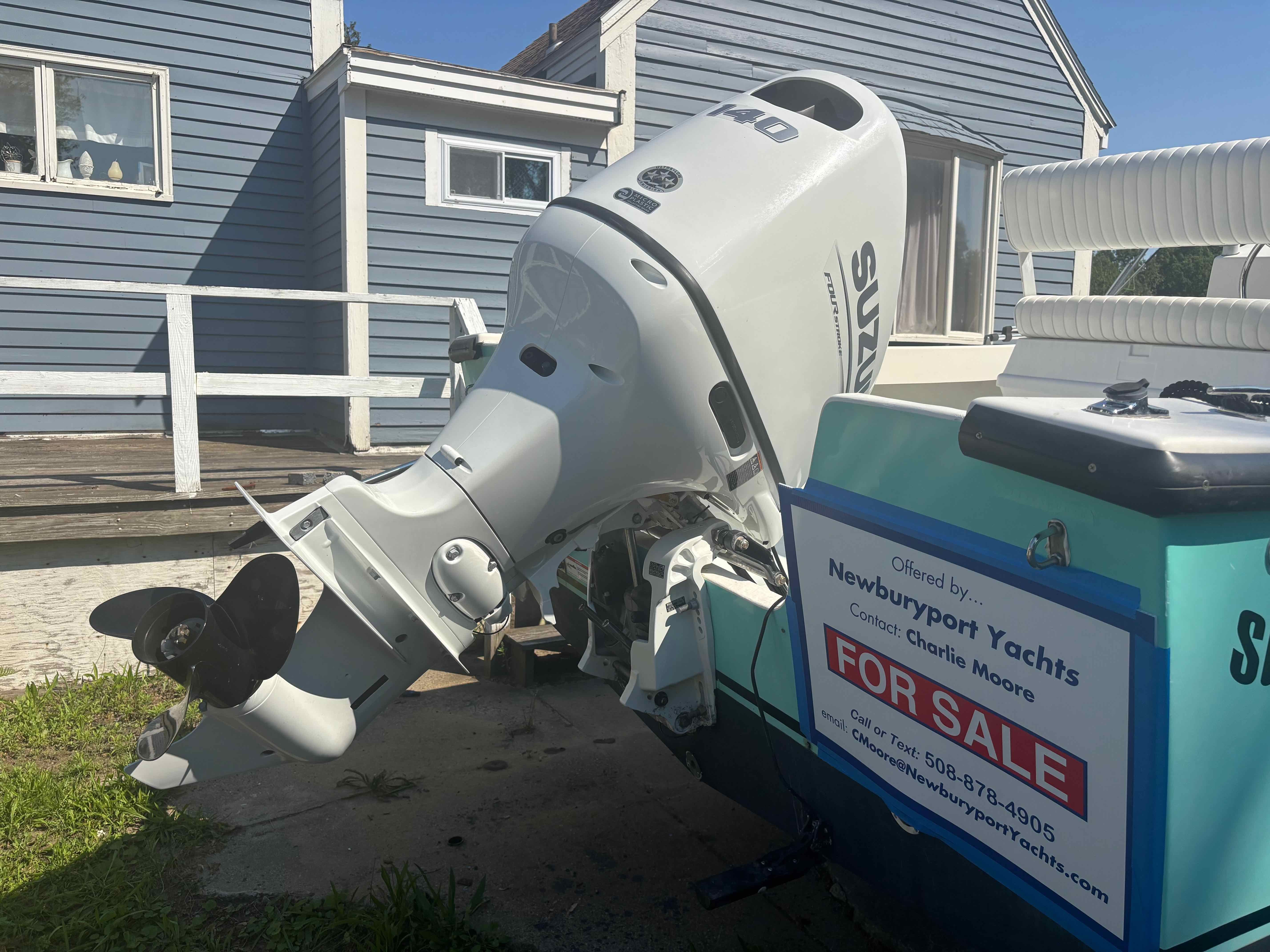 An outboard motor on a boat with a 'For Sale' sign, in front of a blue building.