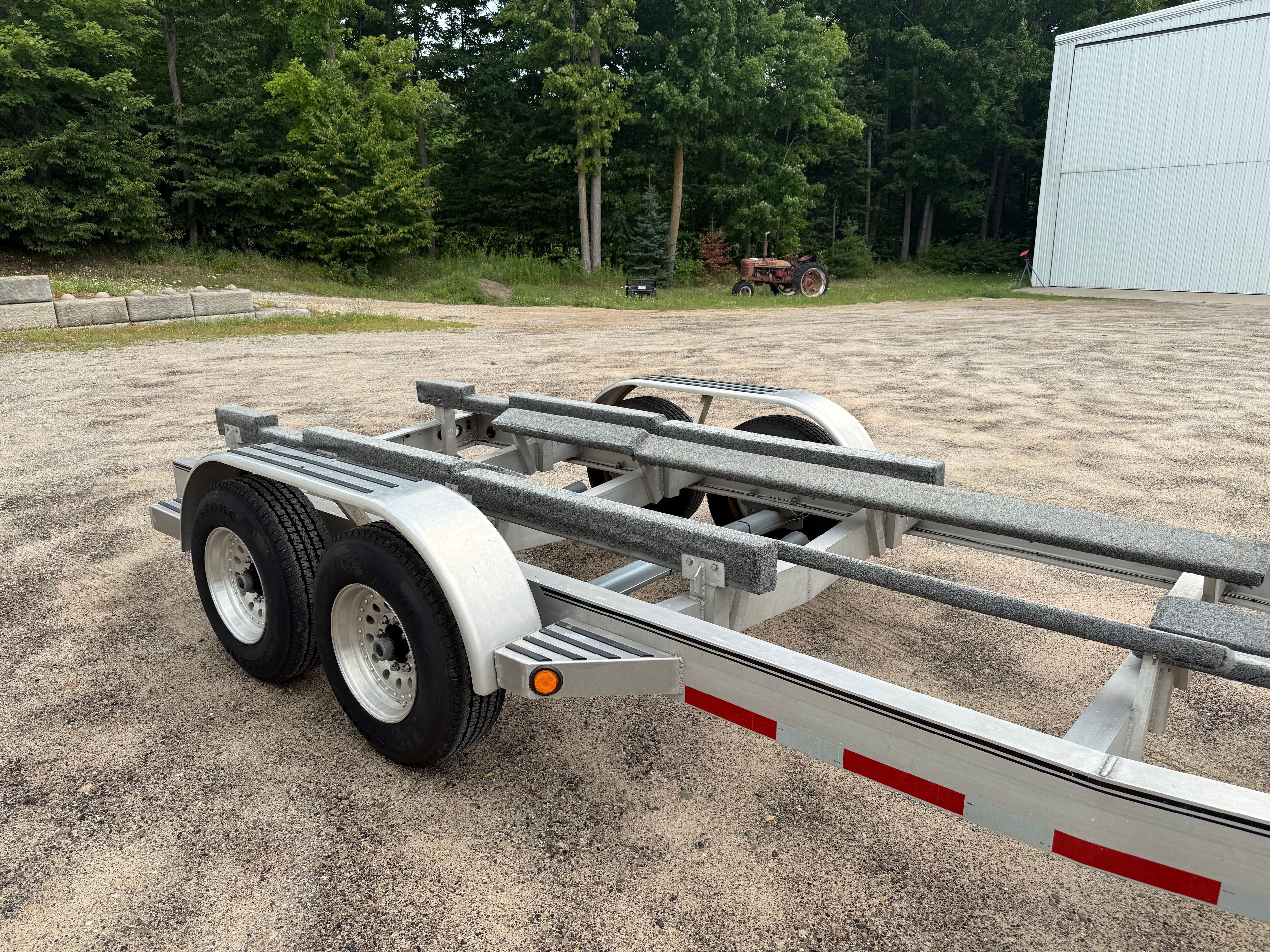 Boat trailer on gravel lot, surrounded by trees and building.