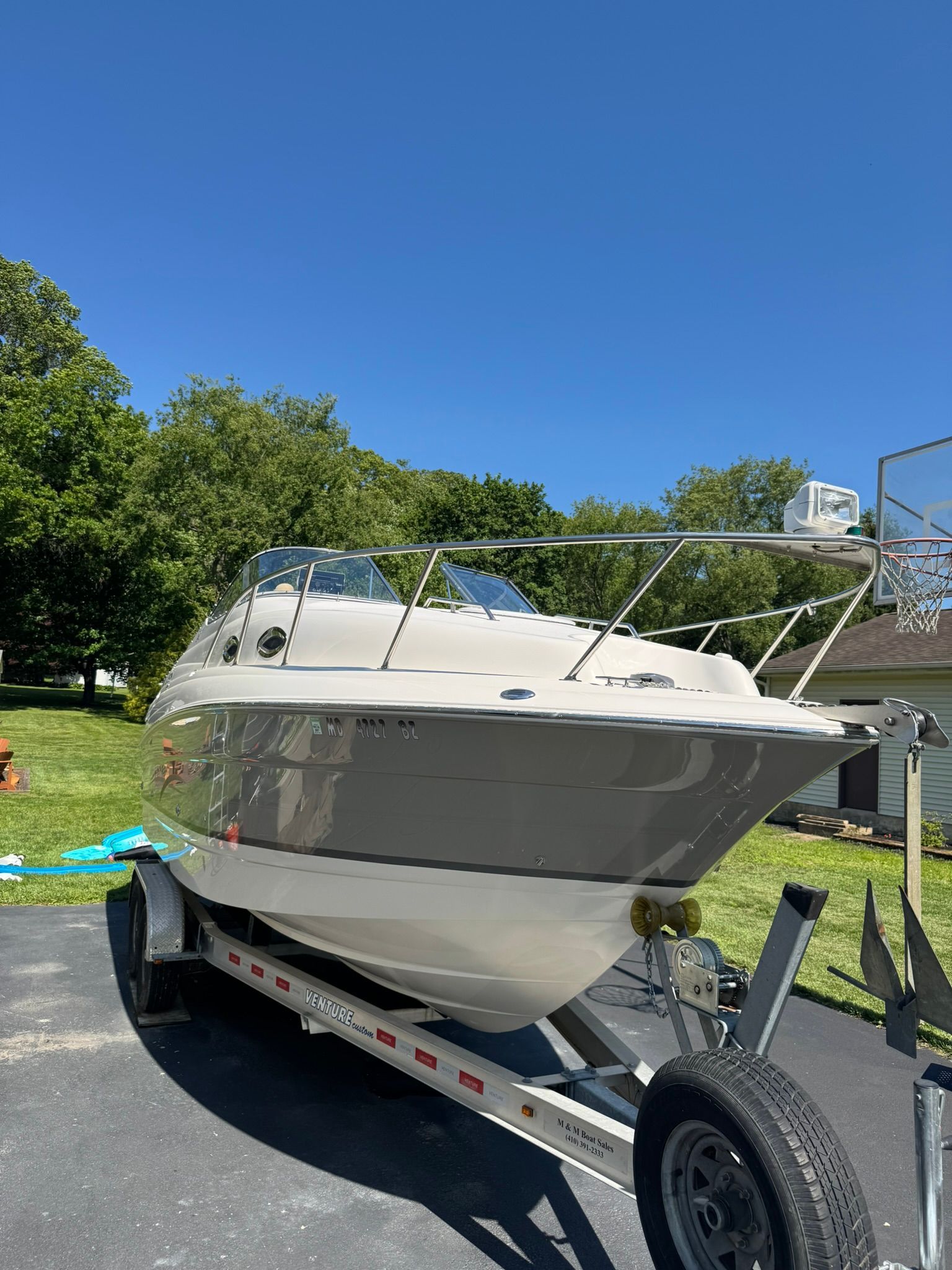 A white motorboat on a trailer is parked on a driveway, surrounded by trees and grass.