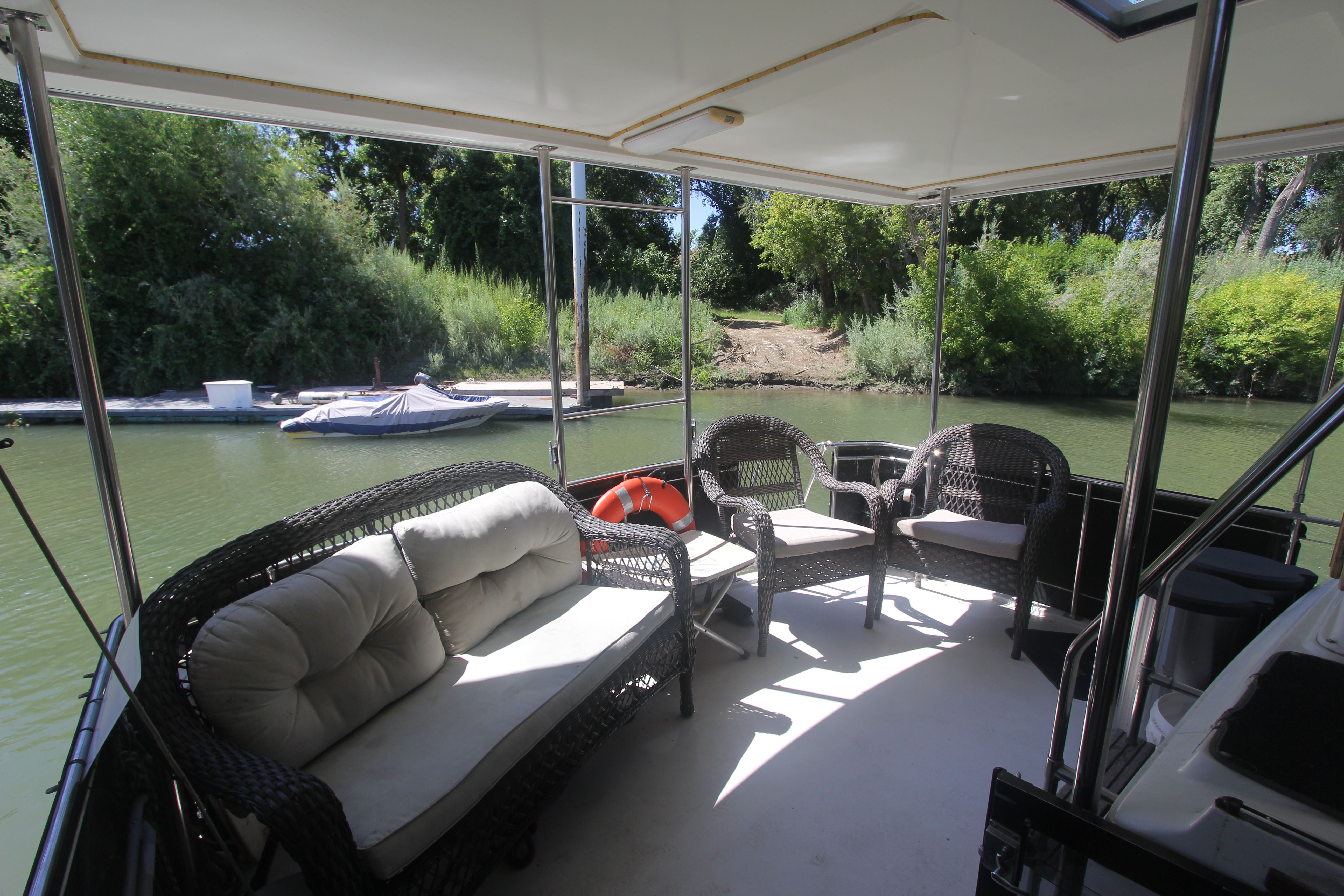 The image shows the aft deck of a 1988 Carver 3607 Aft Cabin Motor Yacht. There are comfortable seating arrangements with wicker furniture and cushions. A calm river with trees is visible in the background.