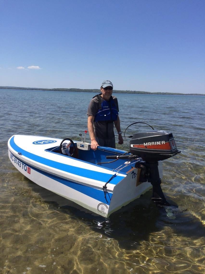 A man stands beside a small blue and white racing boat in shallow water.