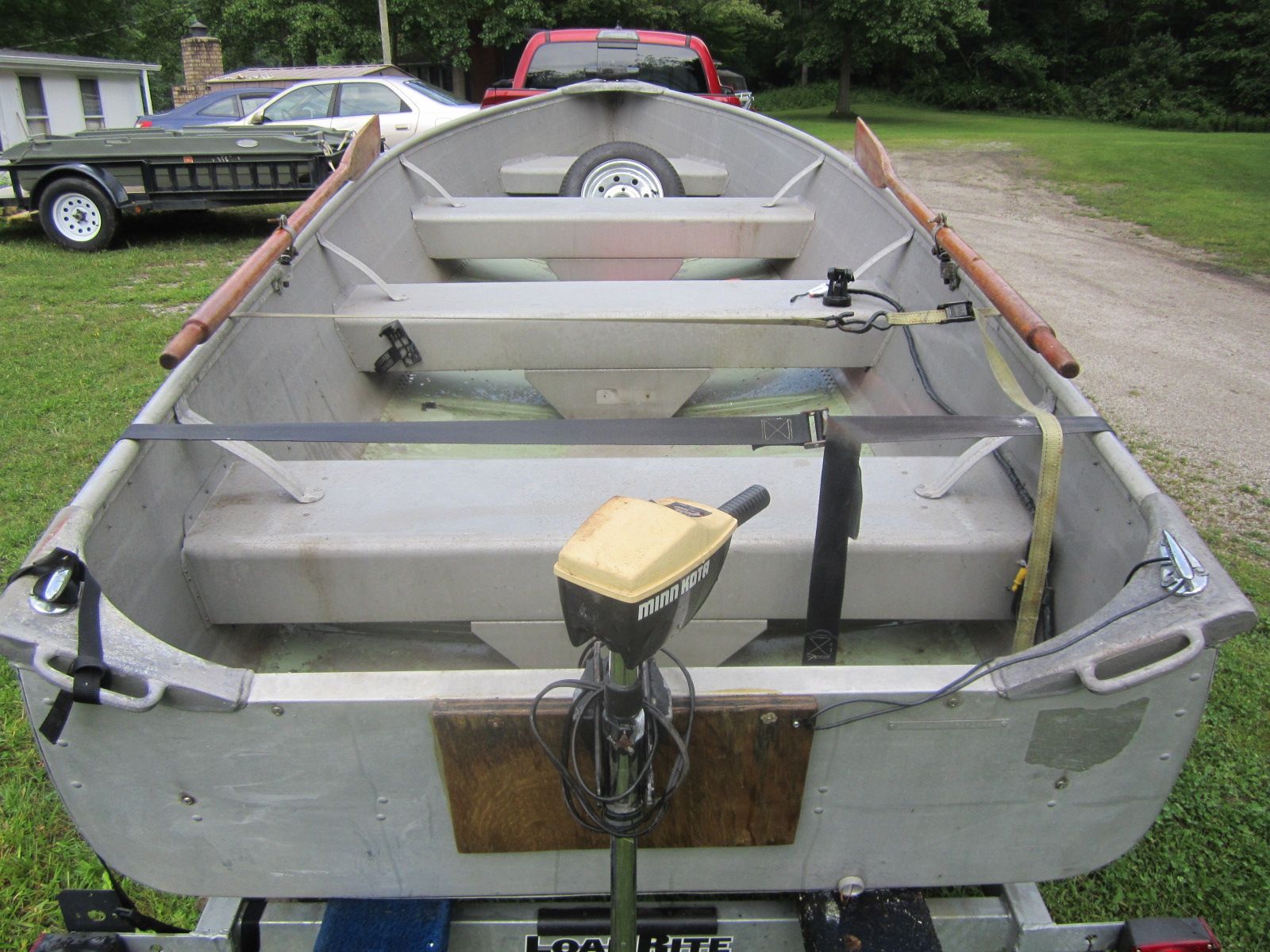 A gray aluminum boat with oars and motor parked on a trailer outdoors.