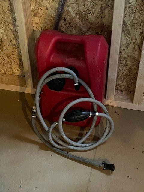 Red fuel tank with hose and pump, leaning against a wooden wall.