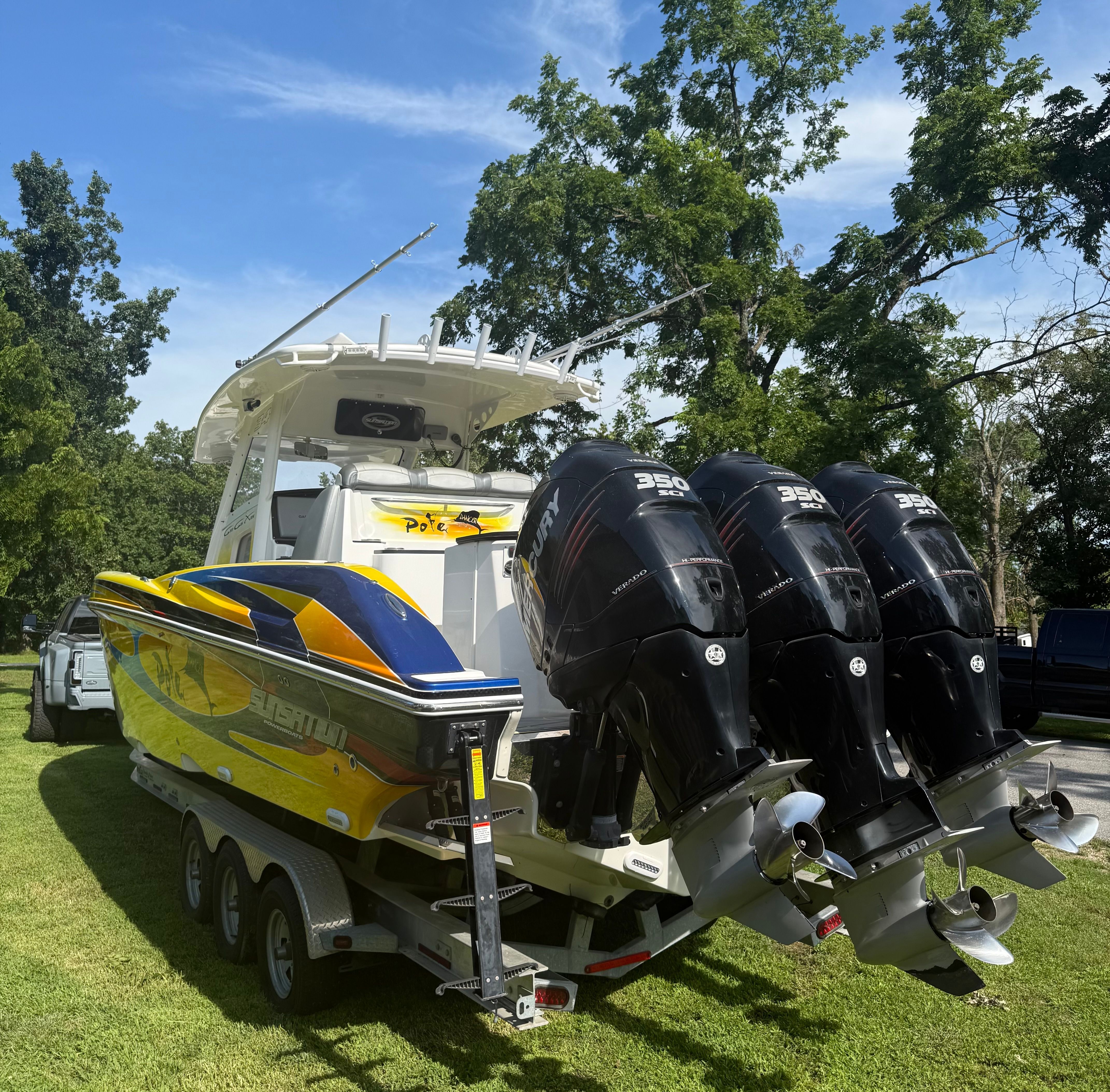 A yellow and blue boat with three outboard engines is on a trailer on a grassy area.