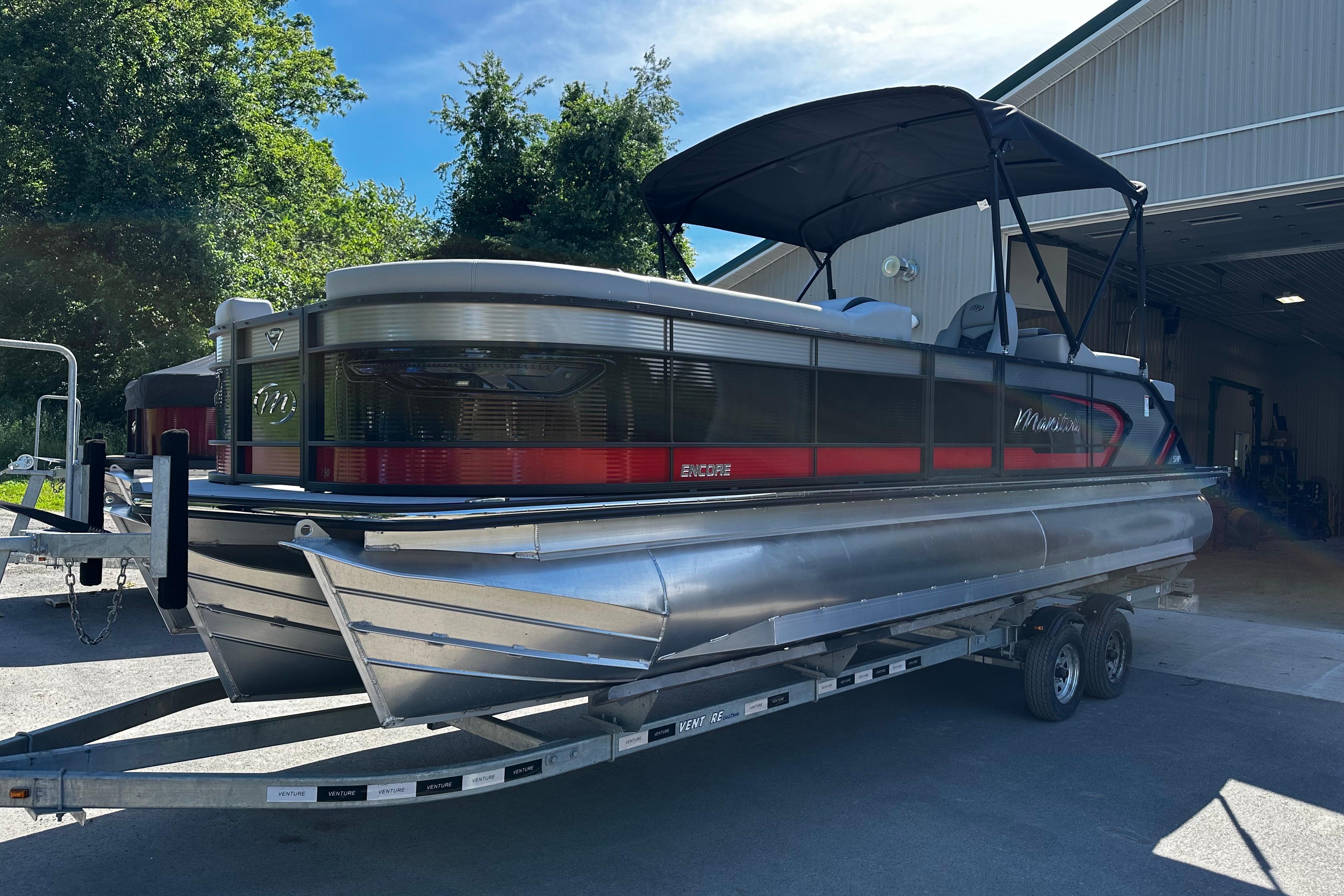 2027 Manitou 25 Encore RF pontoon boat on trailer, parked outside a building.