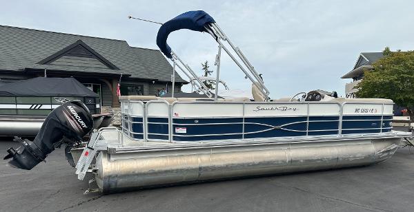 A 2018 South Bay S224CR pontoon boat with a blue canopy and Mercury outboard motor, parked on pavement.