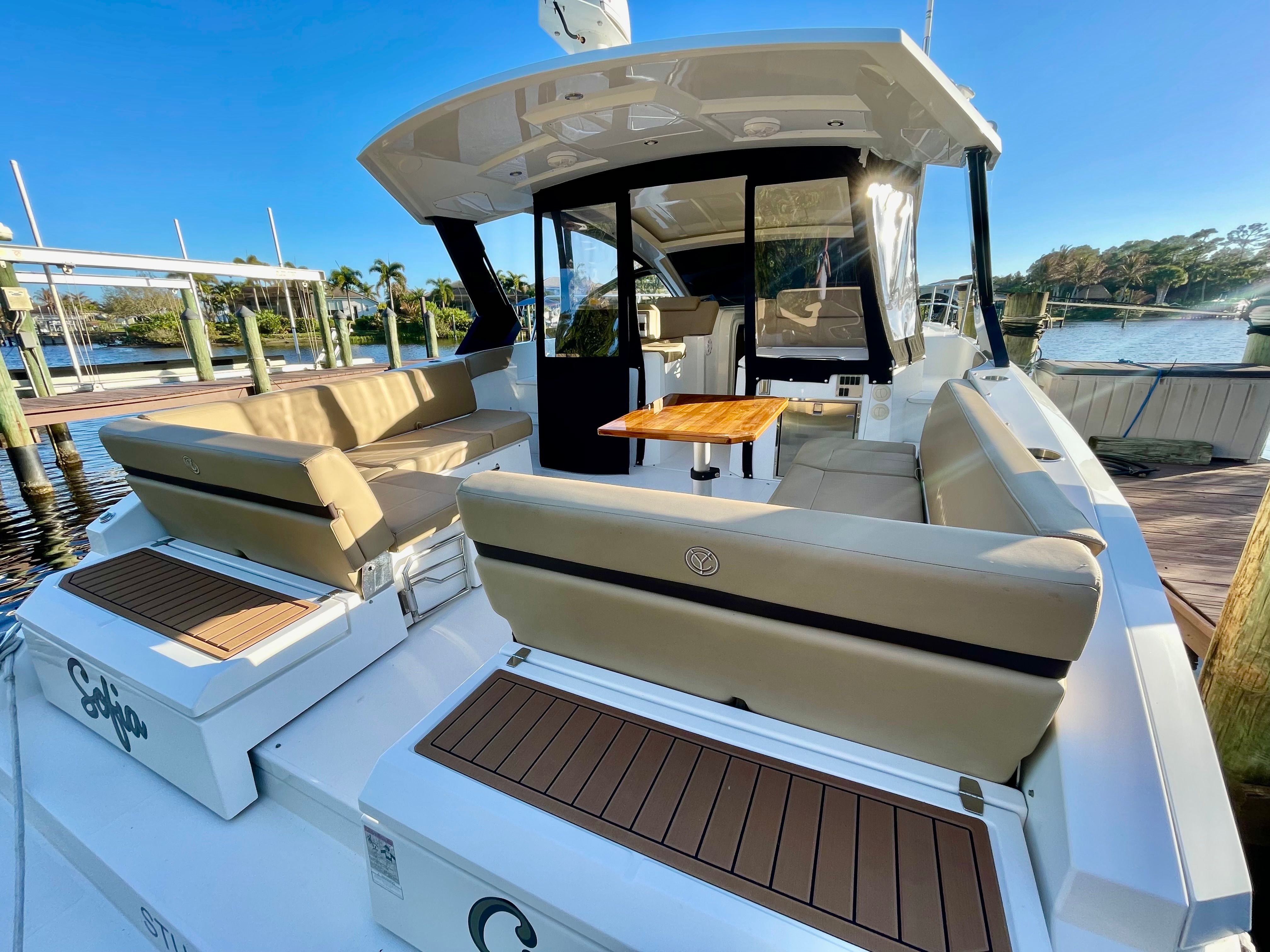 The image shows a luxury yacht with beige seating and a wooden table in an outdoor setting.