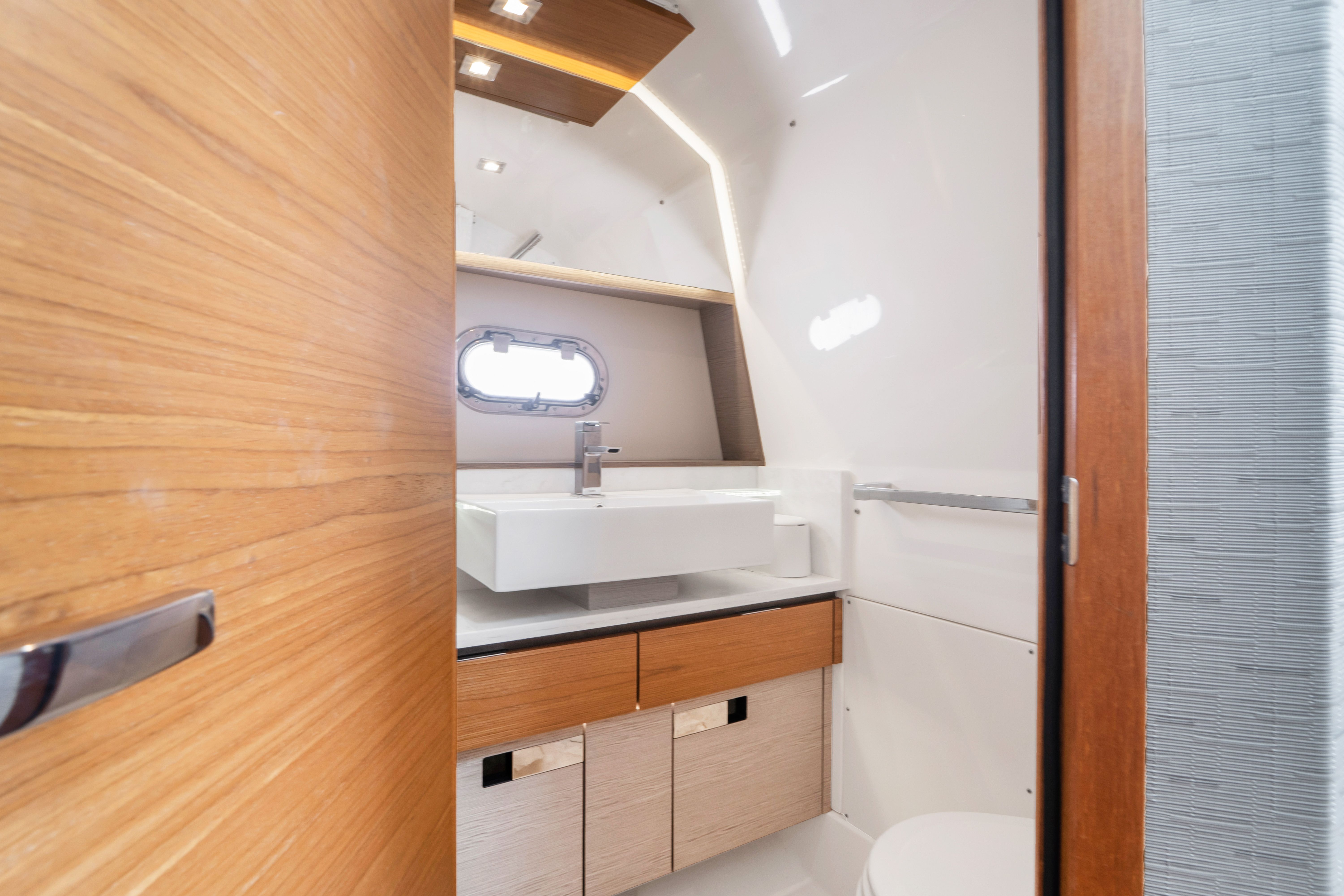 Interior view of a modern boat bathroom with sink, wooden cabinets, and porthole.