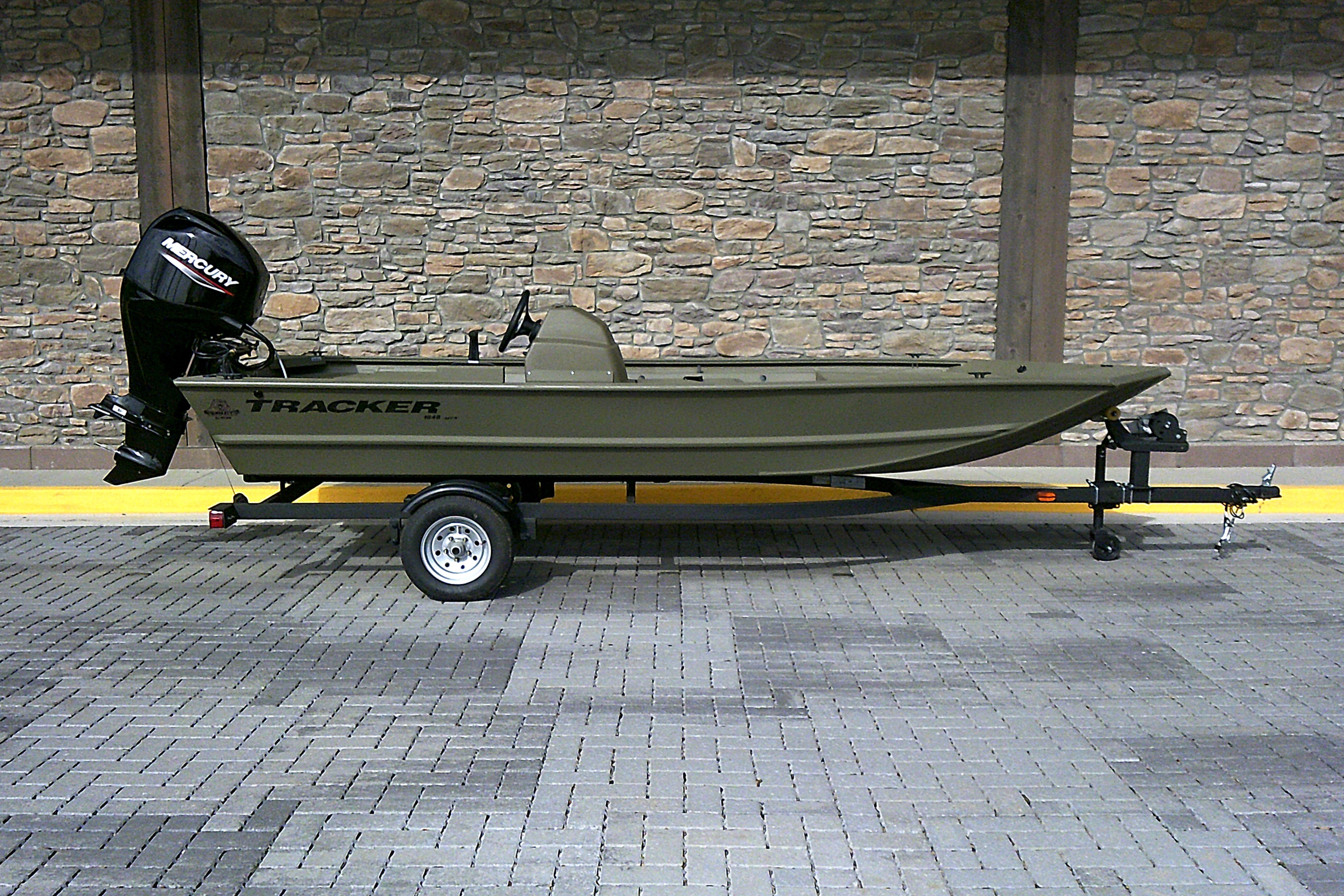 2026 Tracker Grizzly 1648 SC boat on trailer, parked against stone wall background.