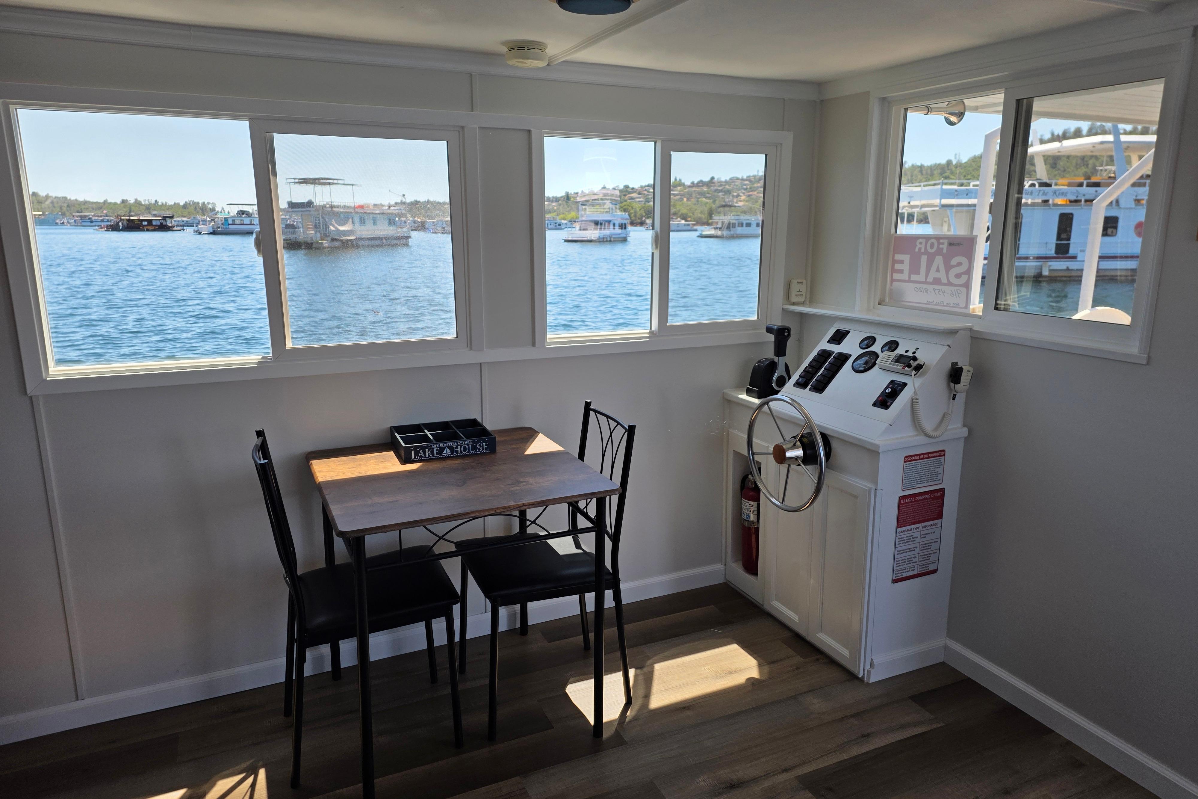 Interior of a 2007 custom houseboat with dining table and helm, overlooking a serene lake.