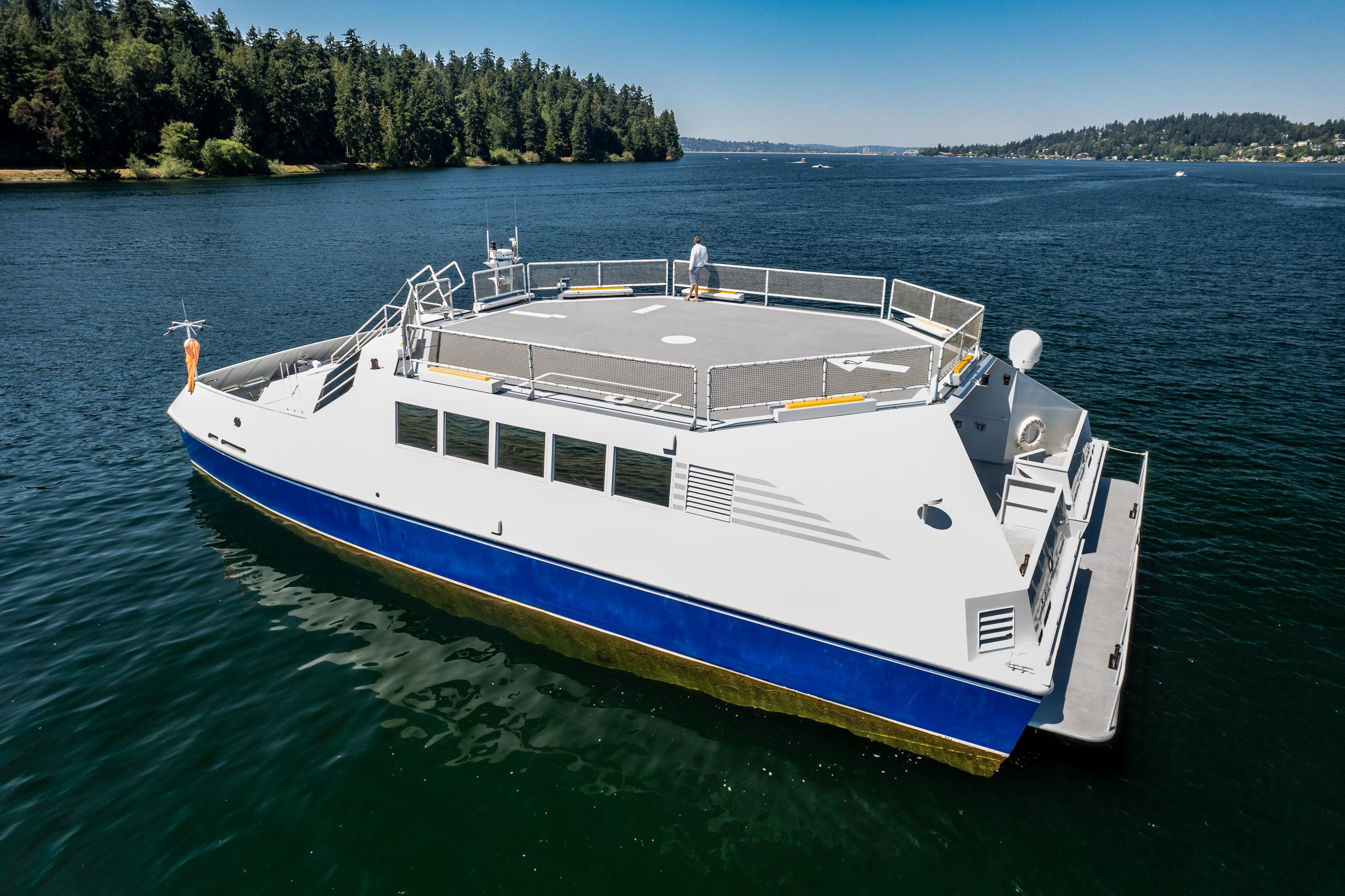 Dakota Creek 1999 catamaran with helicopter landing pad on calm water, surrounded by forested shoreline.