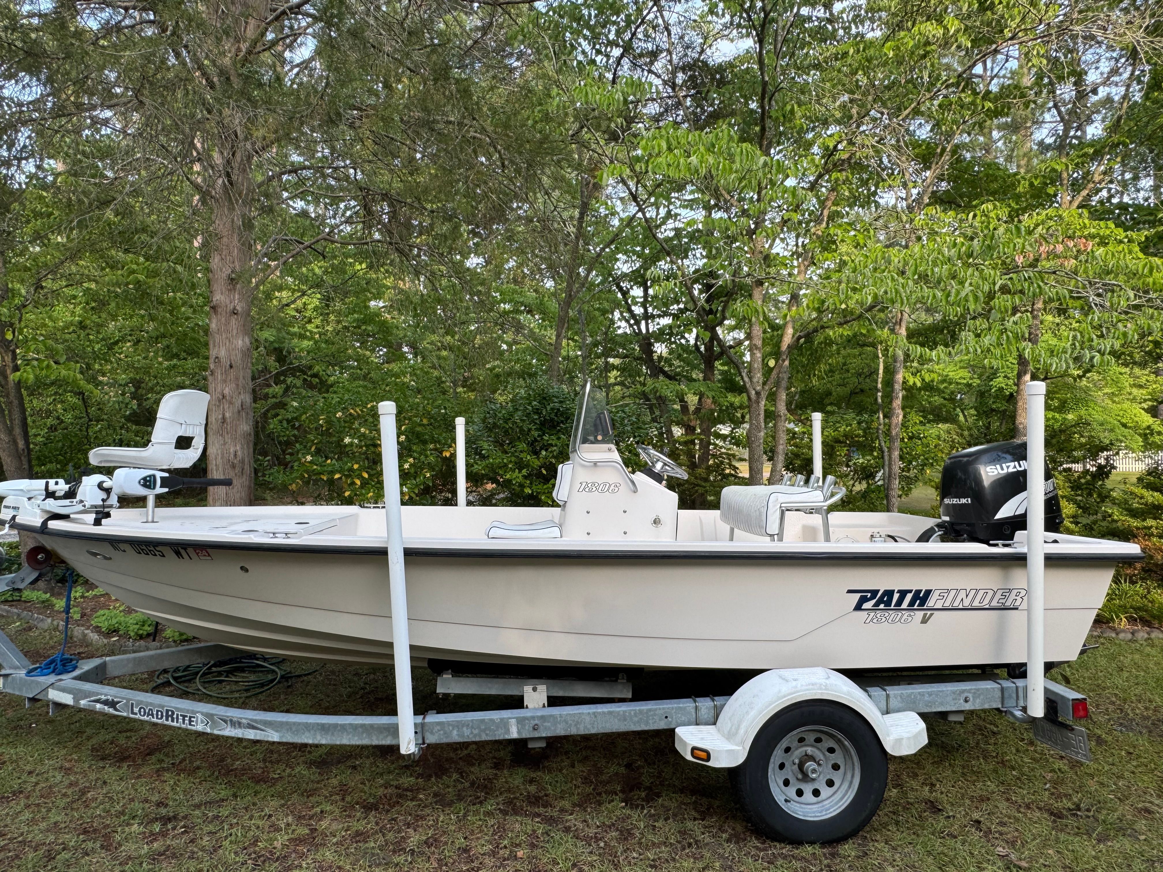 A white boat on a trailer surrounded by trees, featuring an outboard motor.