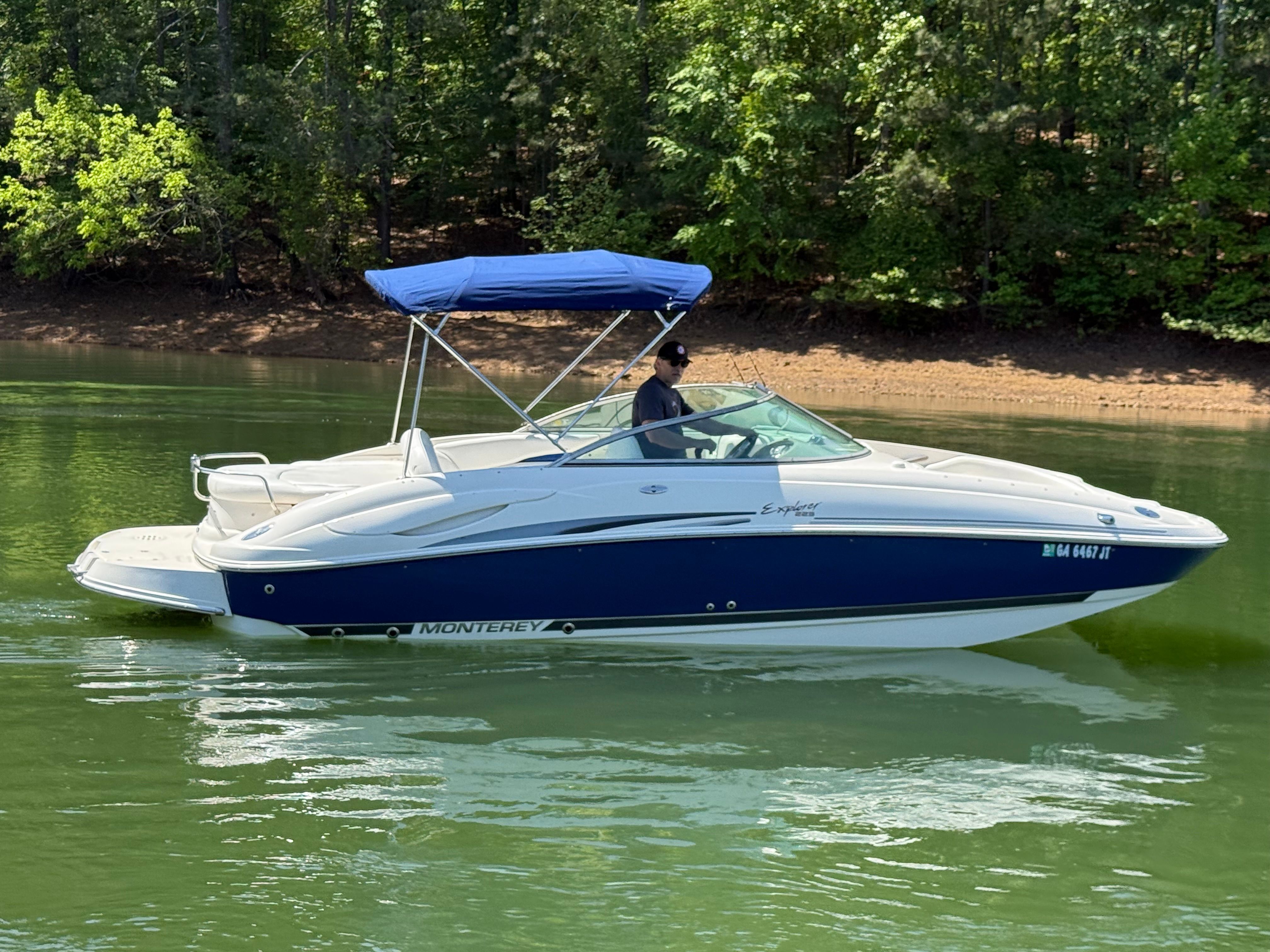 A white and blue 2005 Monterey 223 Explorer Sport boat on a calm lake with a blue sunshade.
