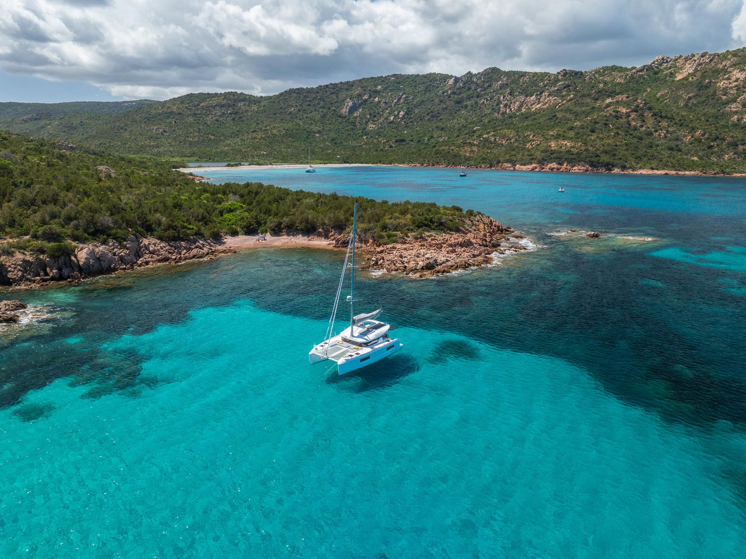 2025 Lagoon 38 catamaran sailing in clear turquoise waters near a rocky coastline.
