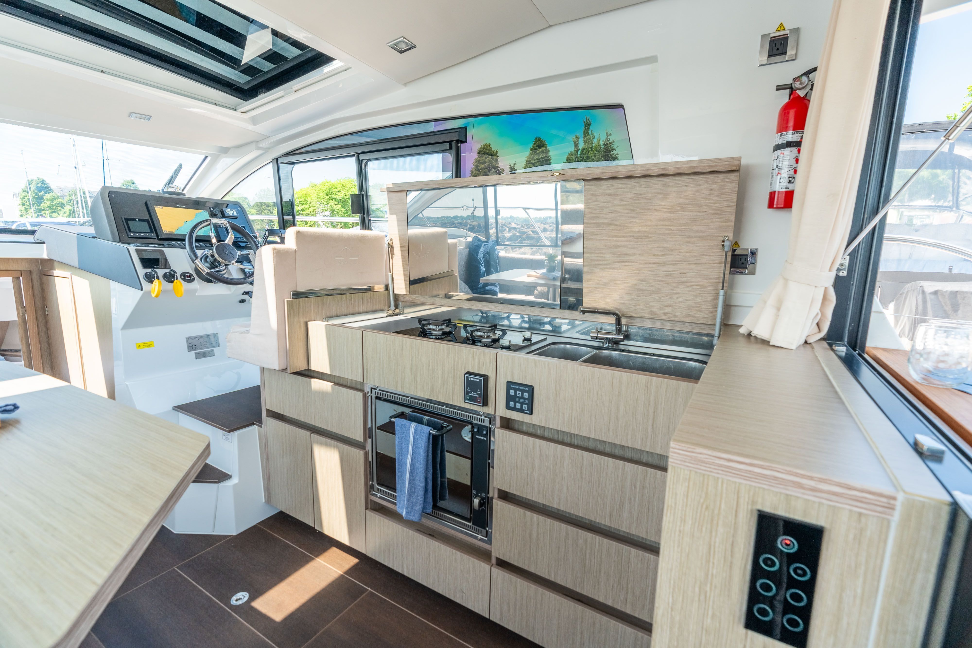 Interior view of a modern boat kitchen with wooden cabinets, stove, and helm station.