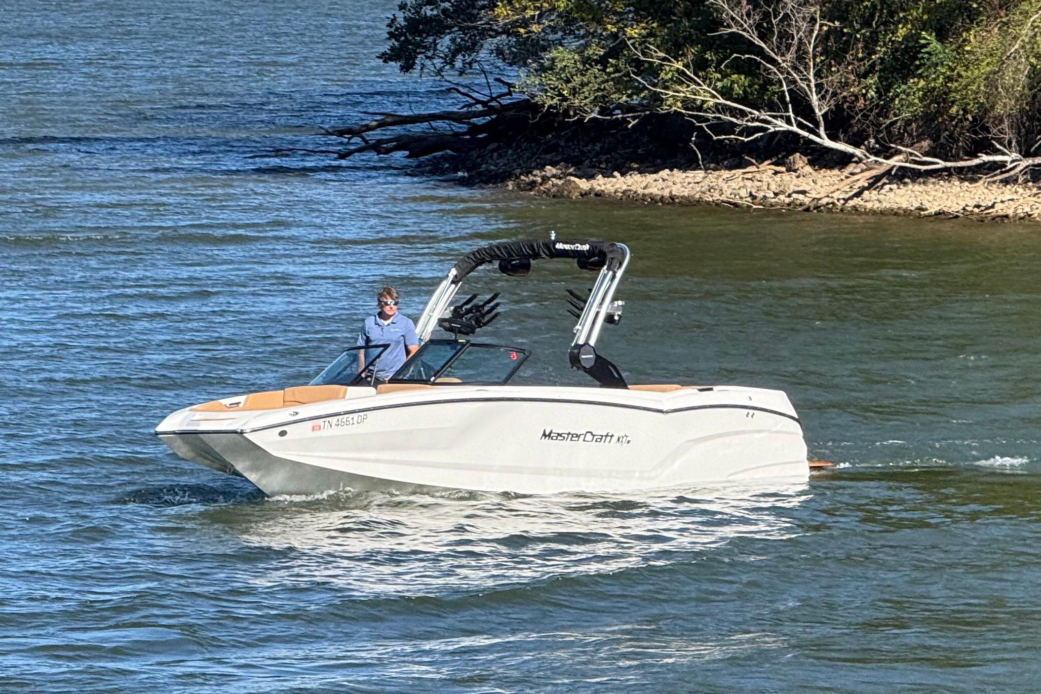 2022 MasterCraft NXT24 boat cruising on a lake near a wooded shoreline.