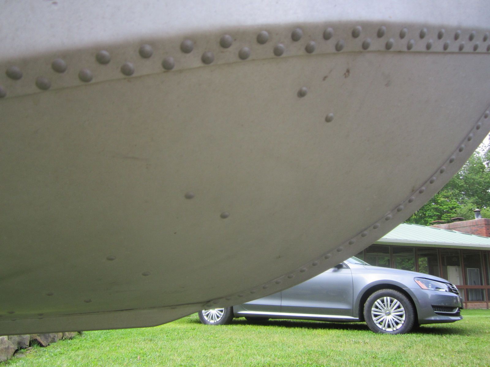 A riveted boat hull beside a car on grass, in front of a building.