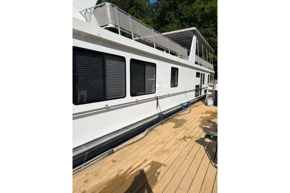 1997 Stardust Houseboat 18x83 with white exterior and wooden deck, docked by lush greenery.