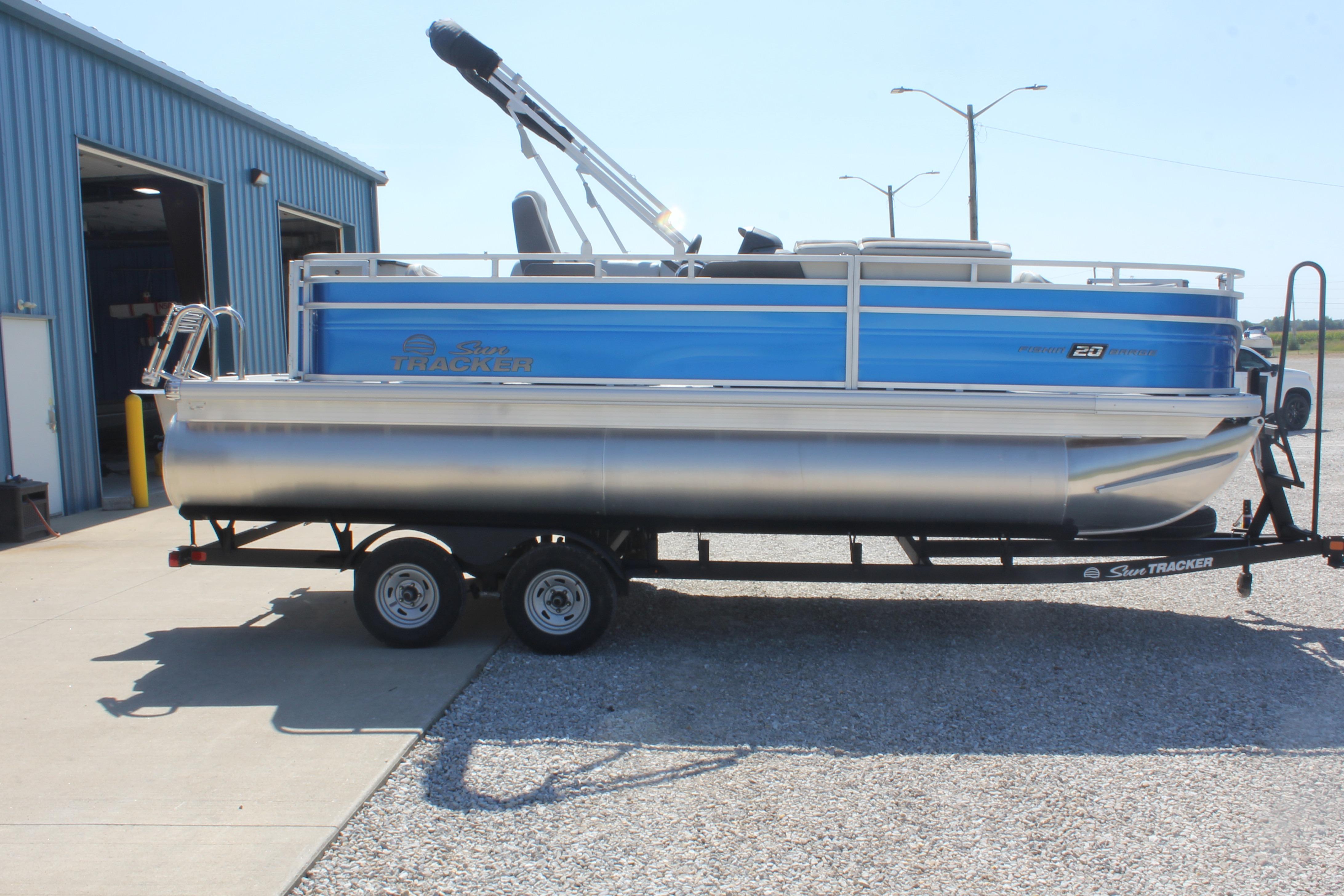 2026 Sun Tracker Fishin' Barge 20 DLX pontoon boat on trailer outside blue building.