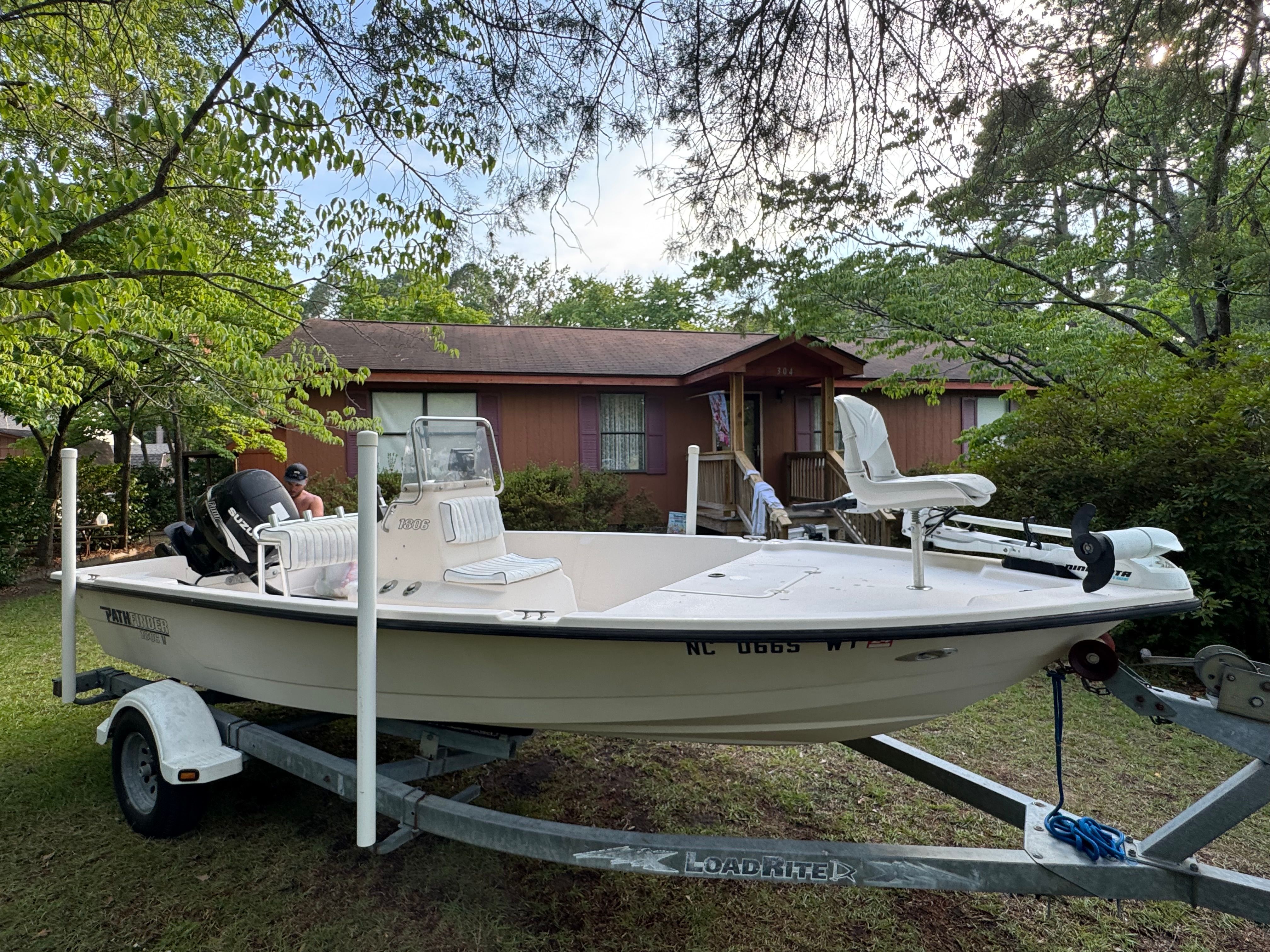 A white boat on a trailer is parked in front of a house, surrounded by trees.