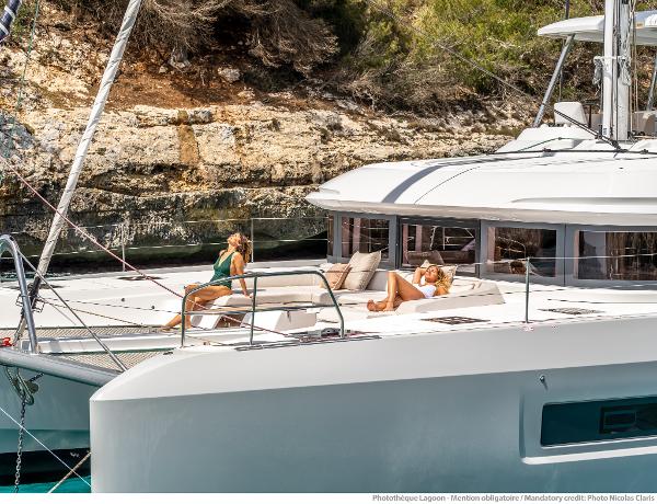 2025 Lagoon 55 yacht with people relaxing on deck, anchored near rocky shoreline.