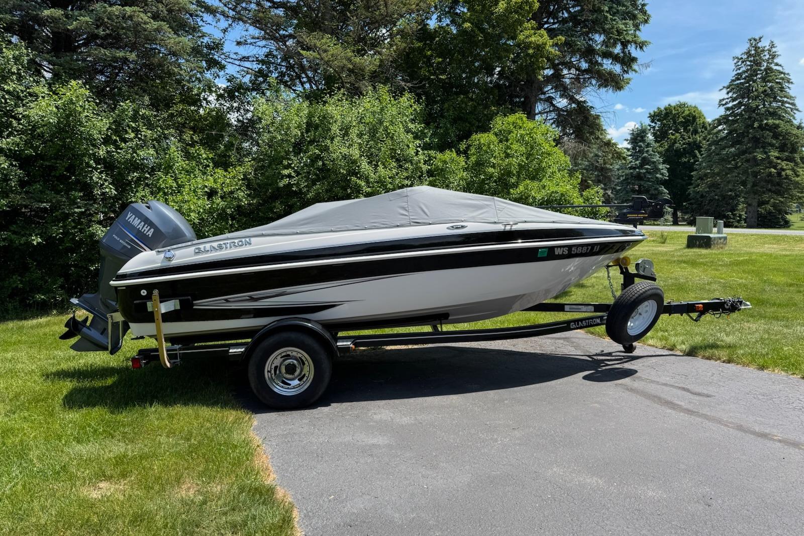 2012 Glastron GT 180 boat on trailer, covered, parked on grass with trees in background.