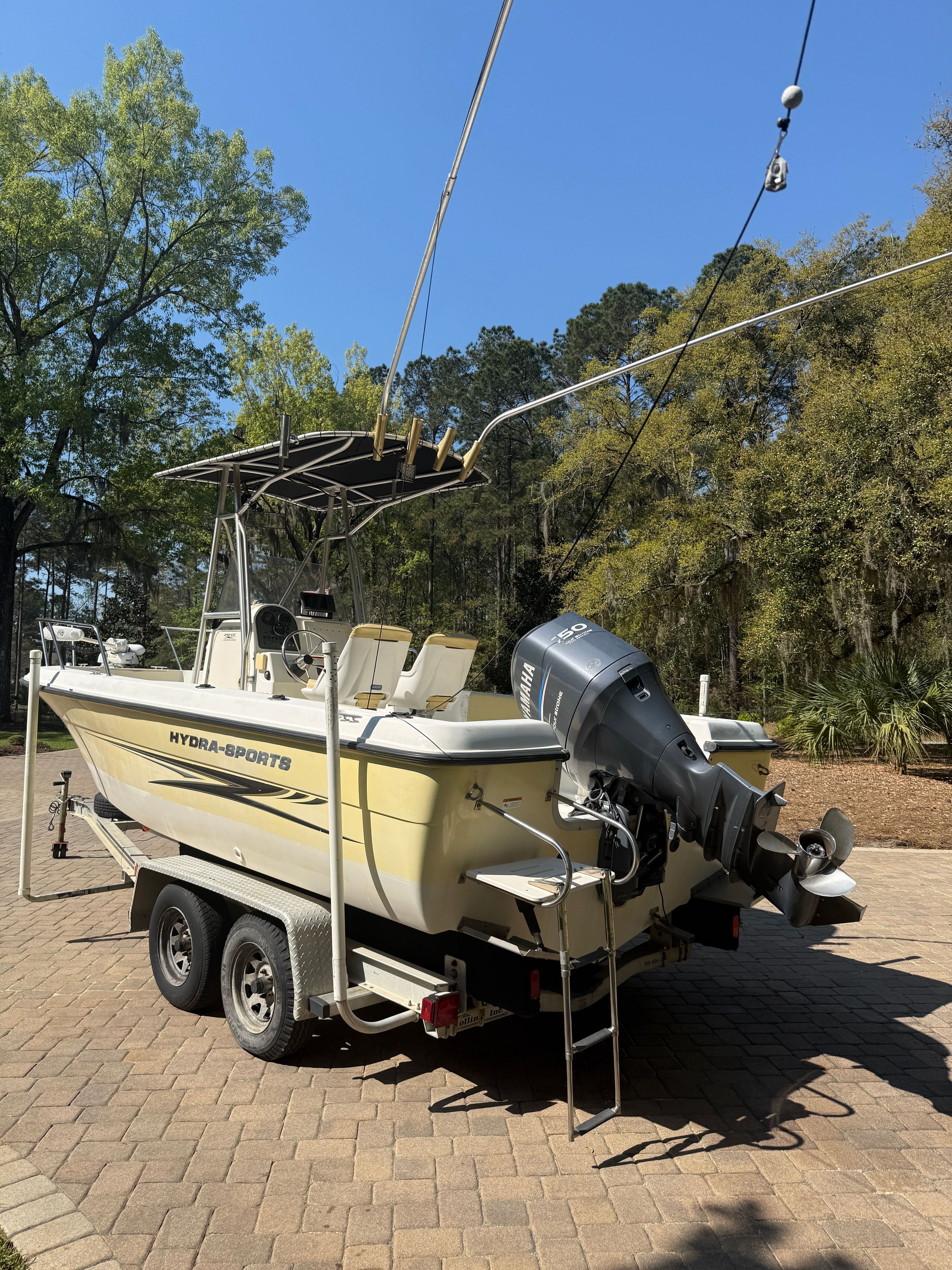 A yellow boat on a trailer, surrounded by trees and a paved ground.