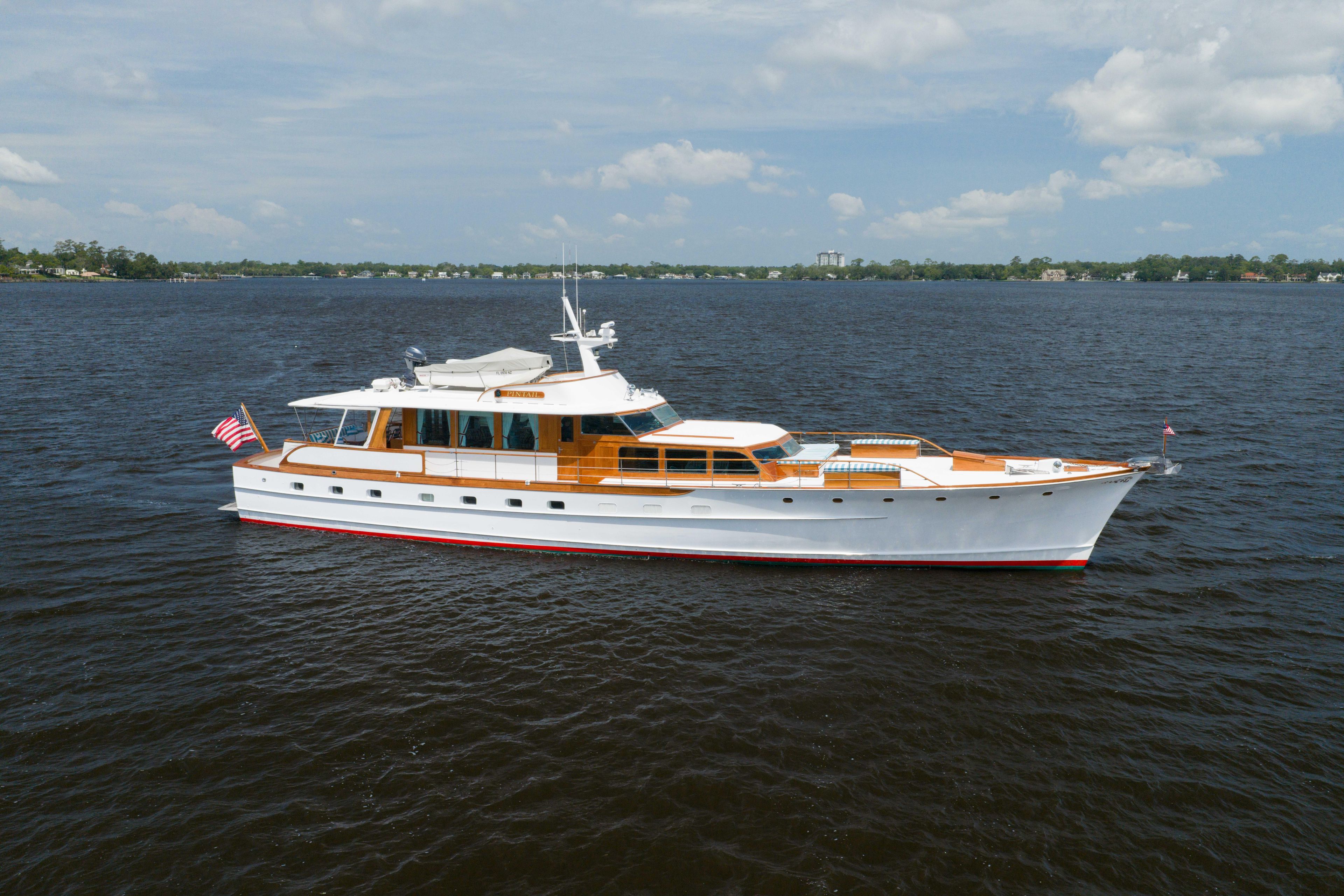 Classic 1963 Trumpy yacht cruising on a serene lake under a partly cloudy sky.