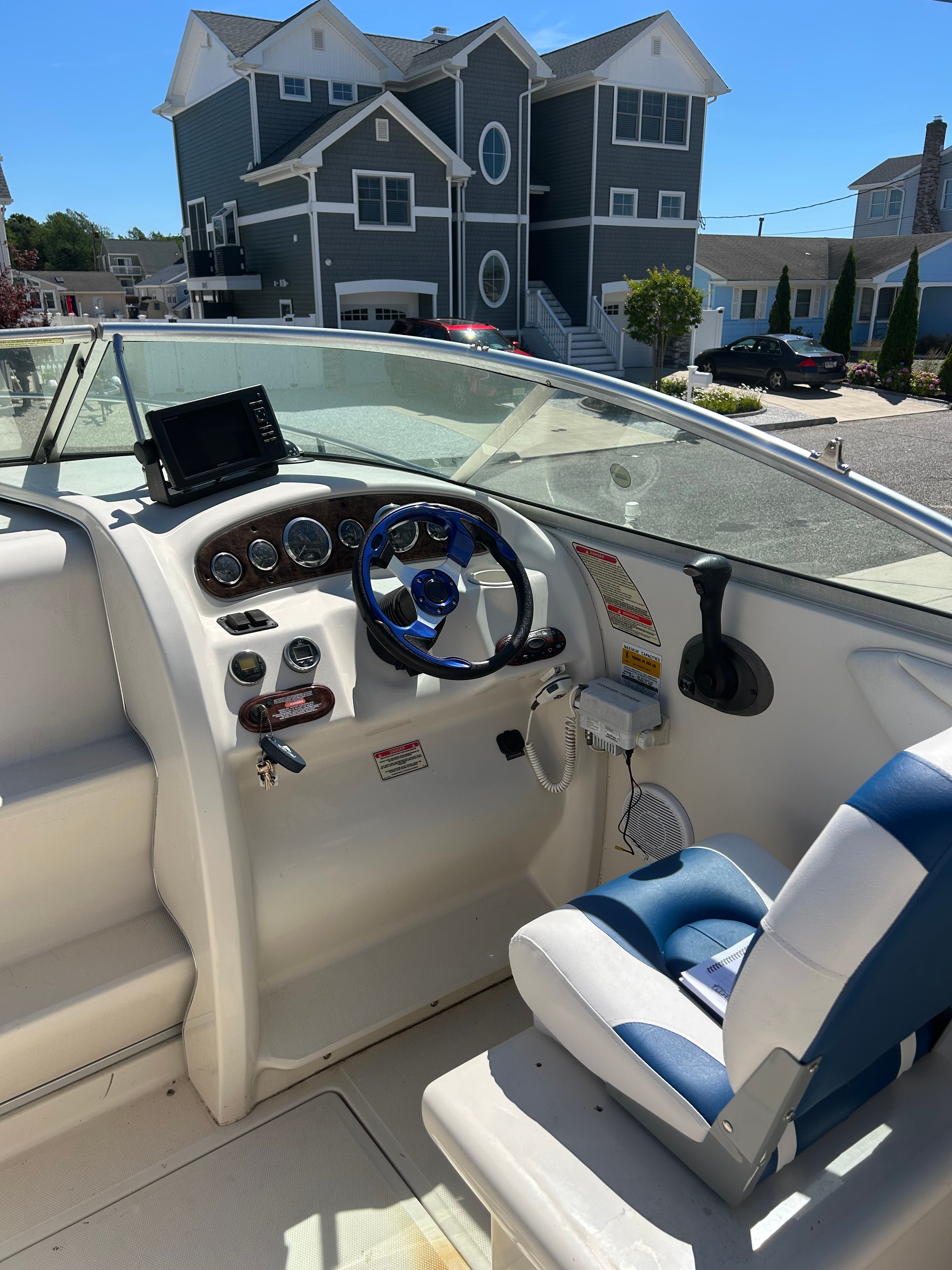 The image shows the cockpit of a 2002 Sea Ray 225 Weekender boat. It features a blue and white steering wheel and navigation system. Houses are visible in the background.