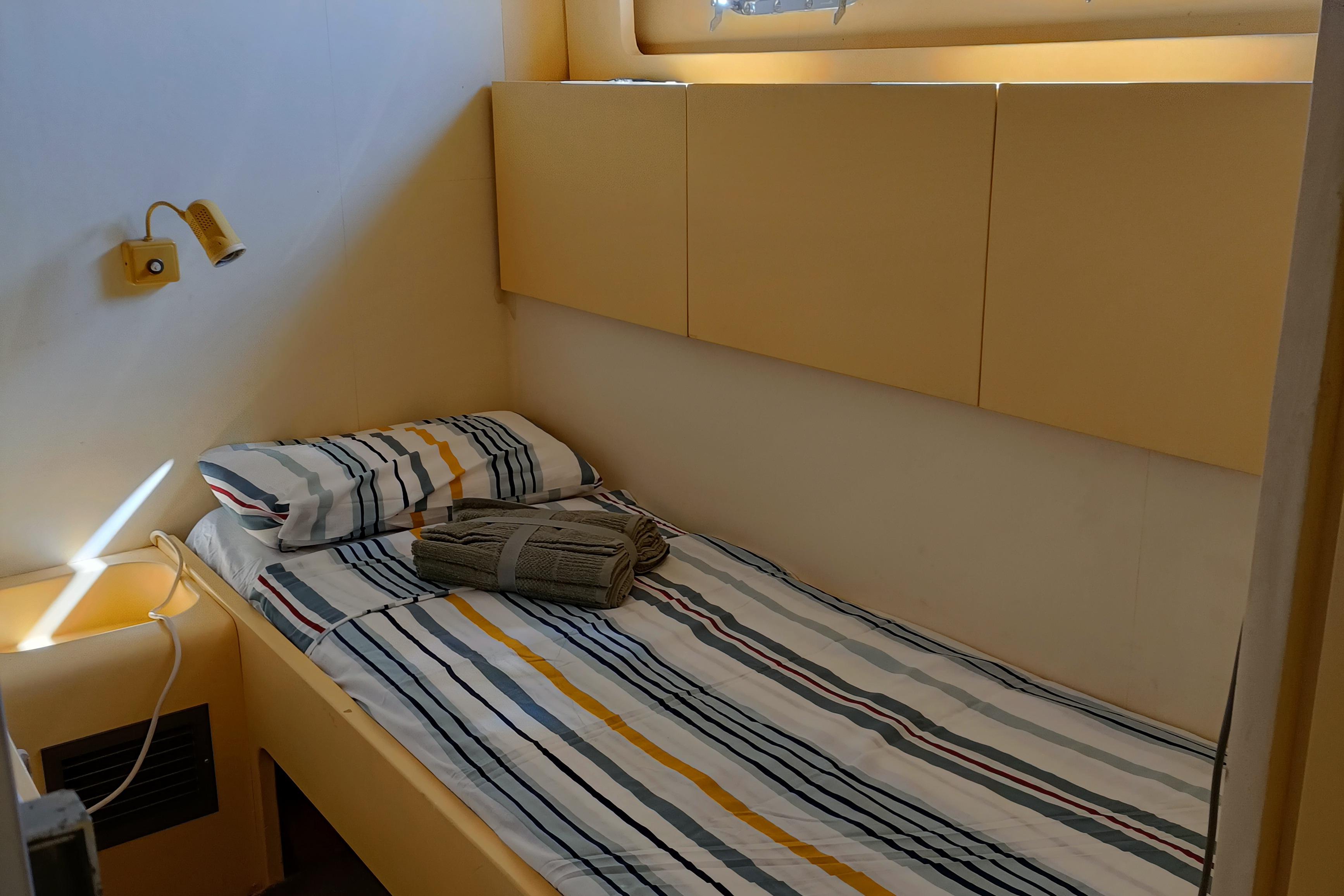Single bed in cozy cabin with striped bedding, yellow walls, and a small reading light. Aresa 16, 1980.