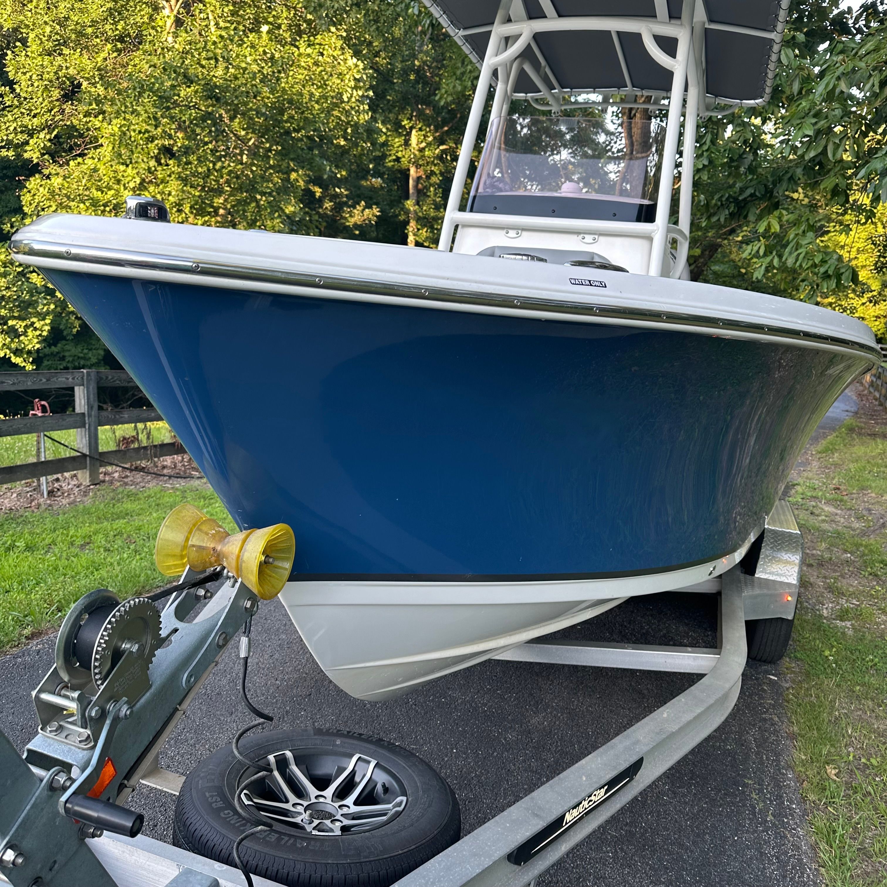A blue 2022 NauticStar 2102 Legacy boat on a trailer, surrounded by trees.