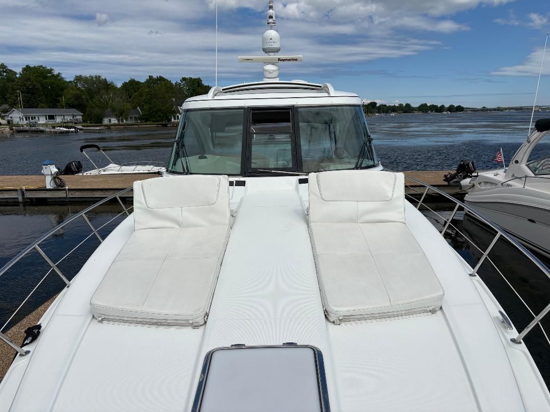 The image shows the foredeck of a white yacht with two cushioned sunbeds. A calm harbor is in the background.