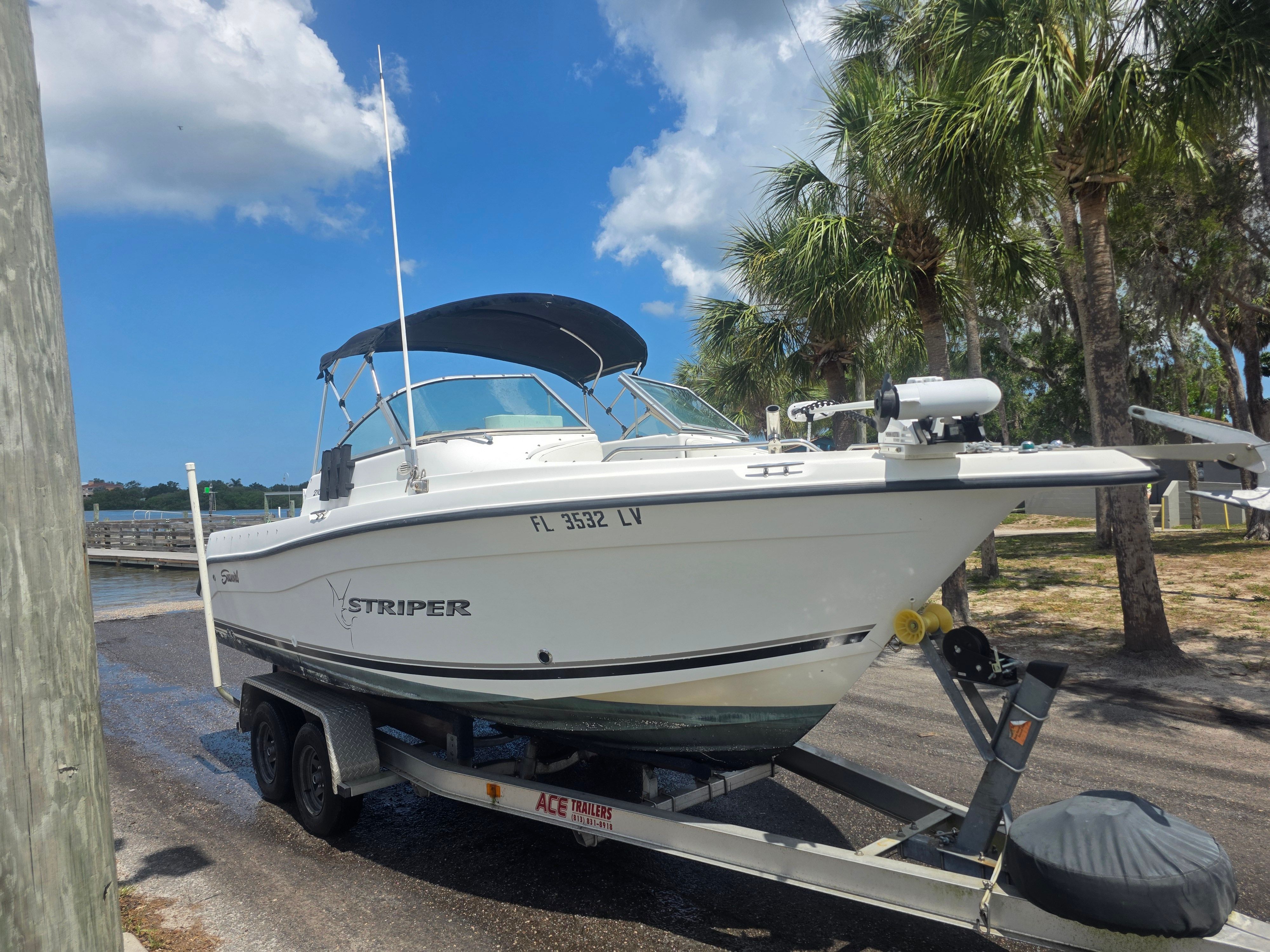 A white boat on a trailer on land, surrounded by palm trees.