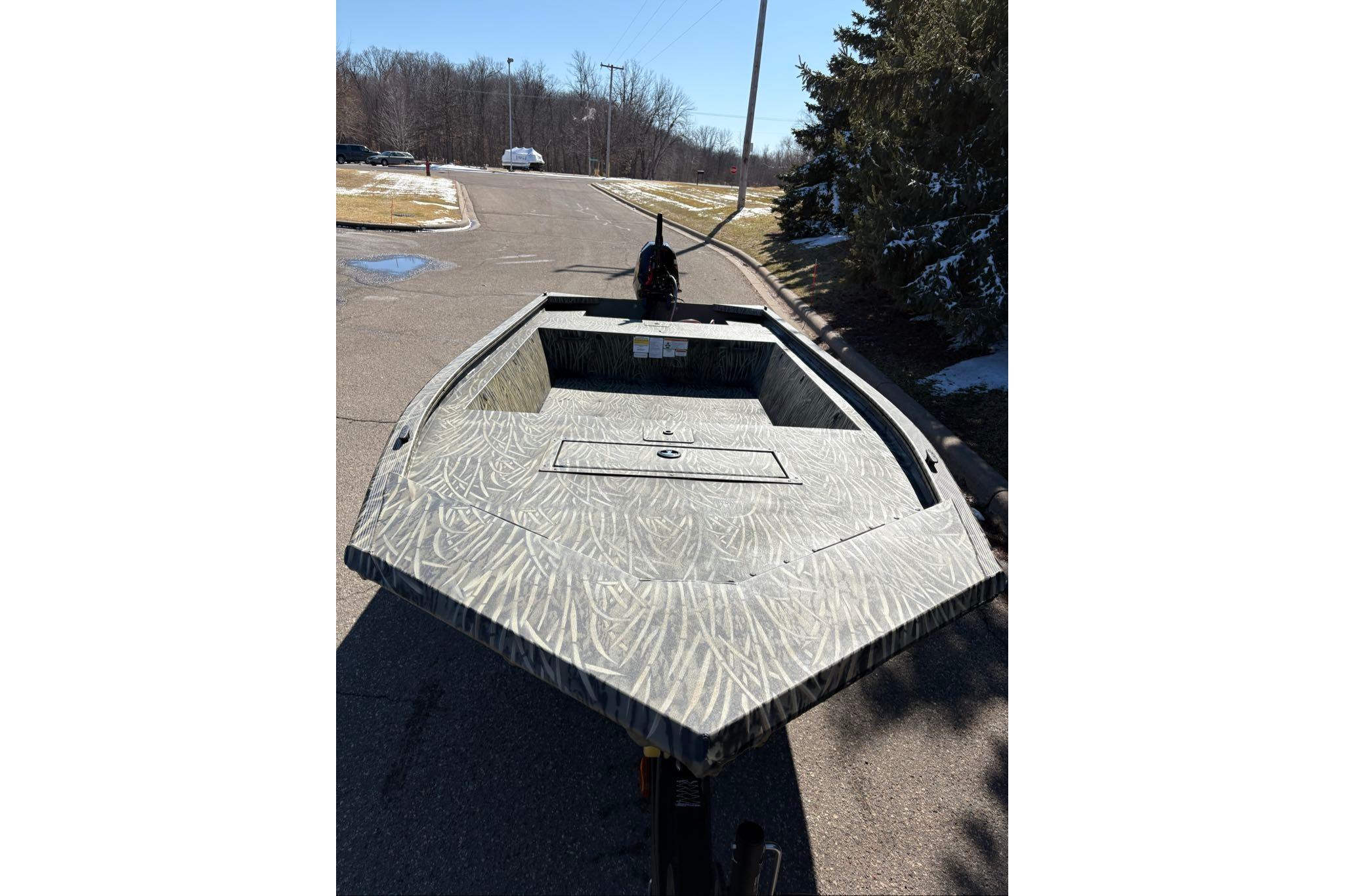 2026 Tracker Grizzly 1648 Utility boat on a paved road, surrounded by trees.