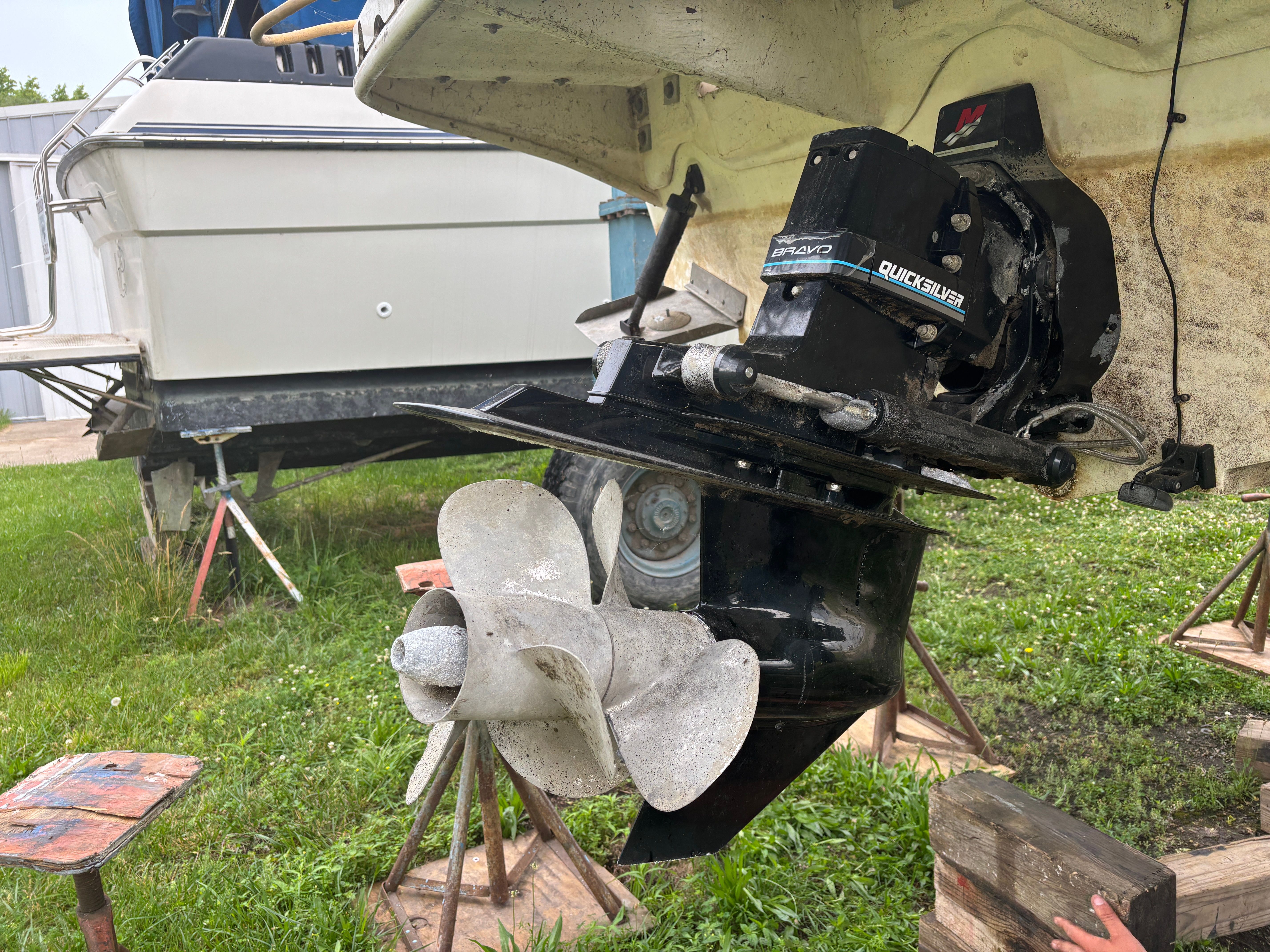 A boat engine with propeller is propped up, surrounded by grass and another boat in the background.