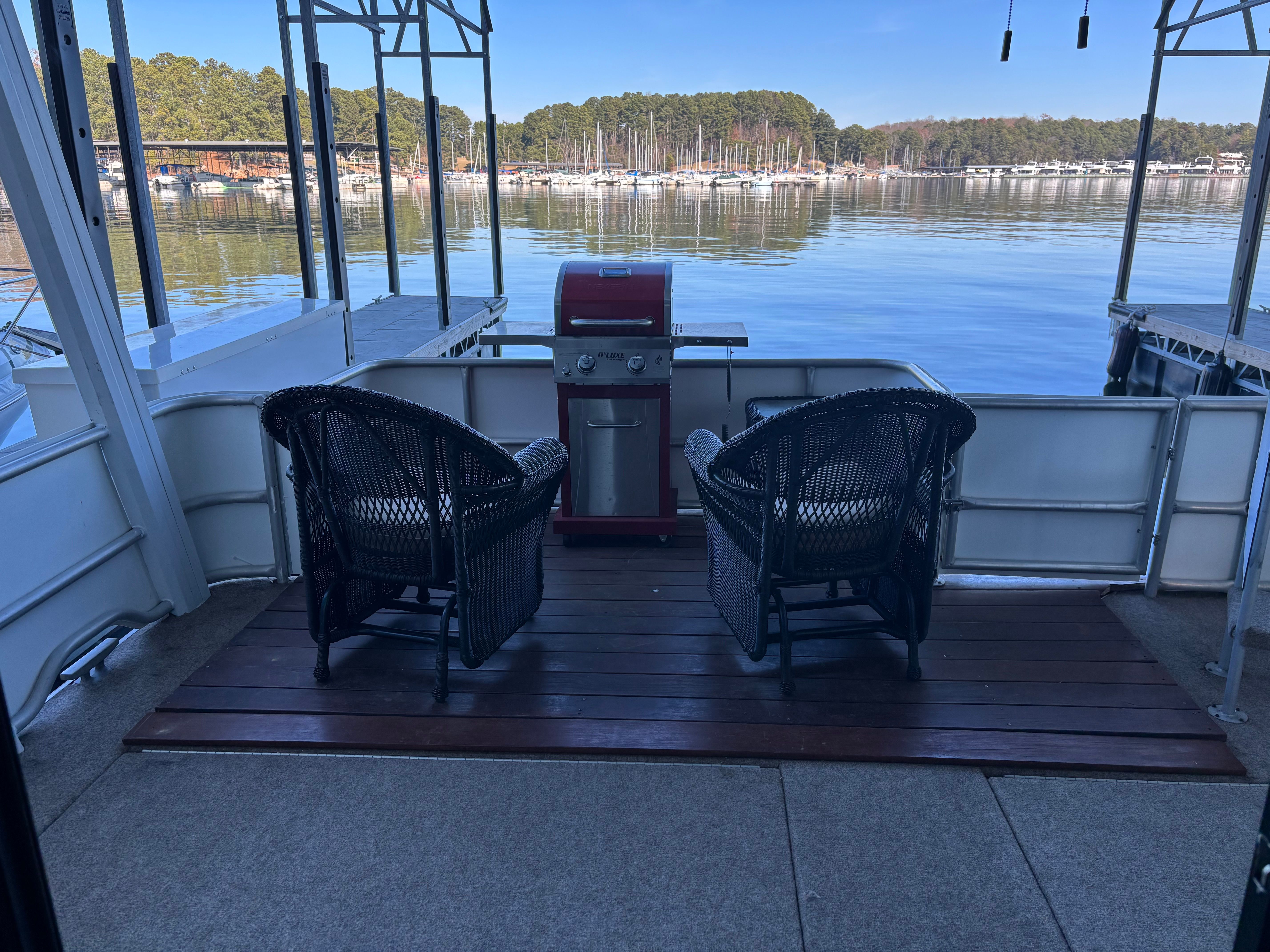Two wicker chairs on a boat with a grill, overlooking a calm lake and forest.
