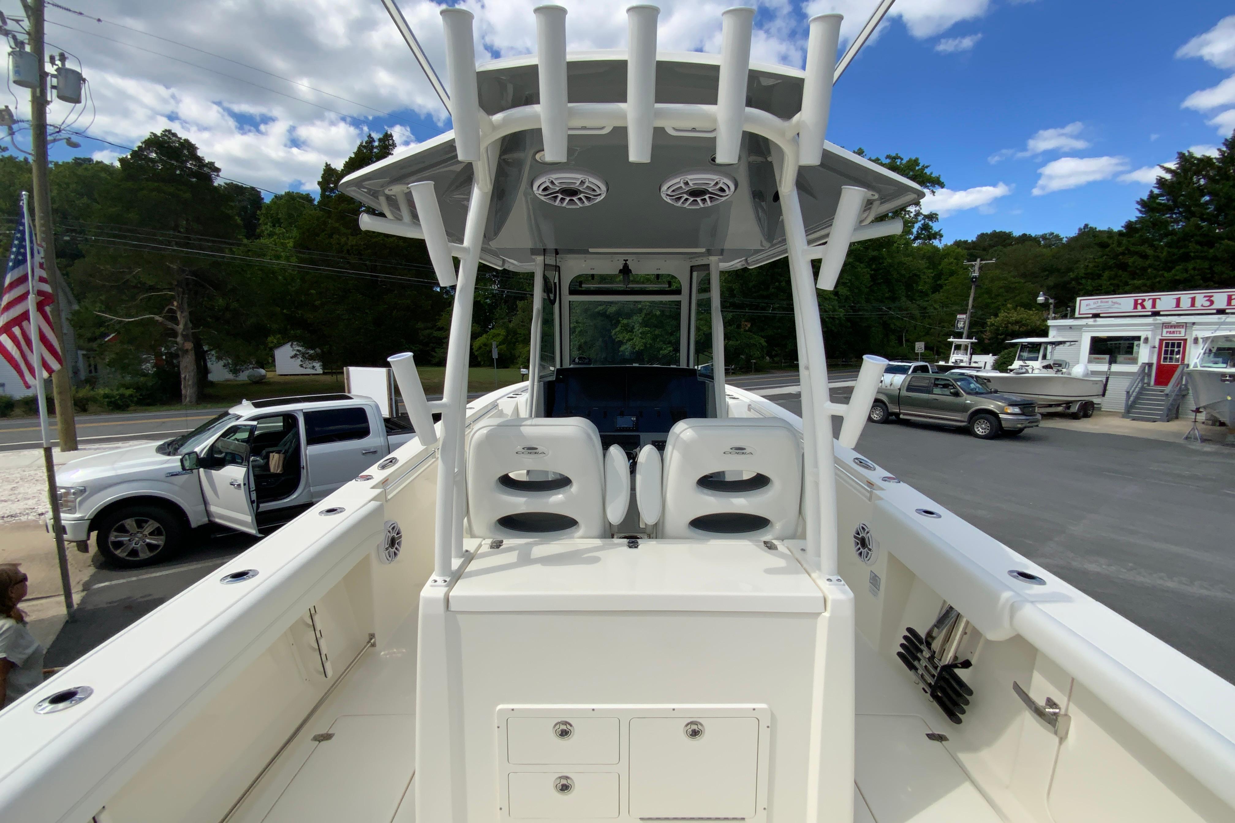 2027 Cobia 320 Center Console boat with T-top, docked near vehicles and trees.