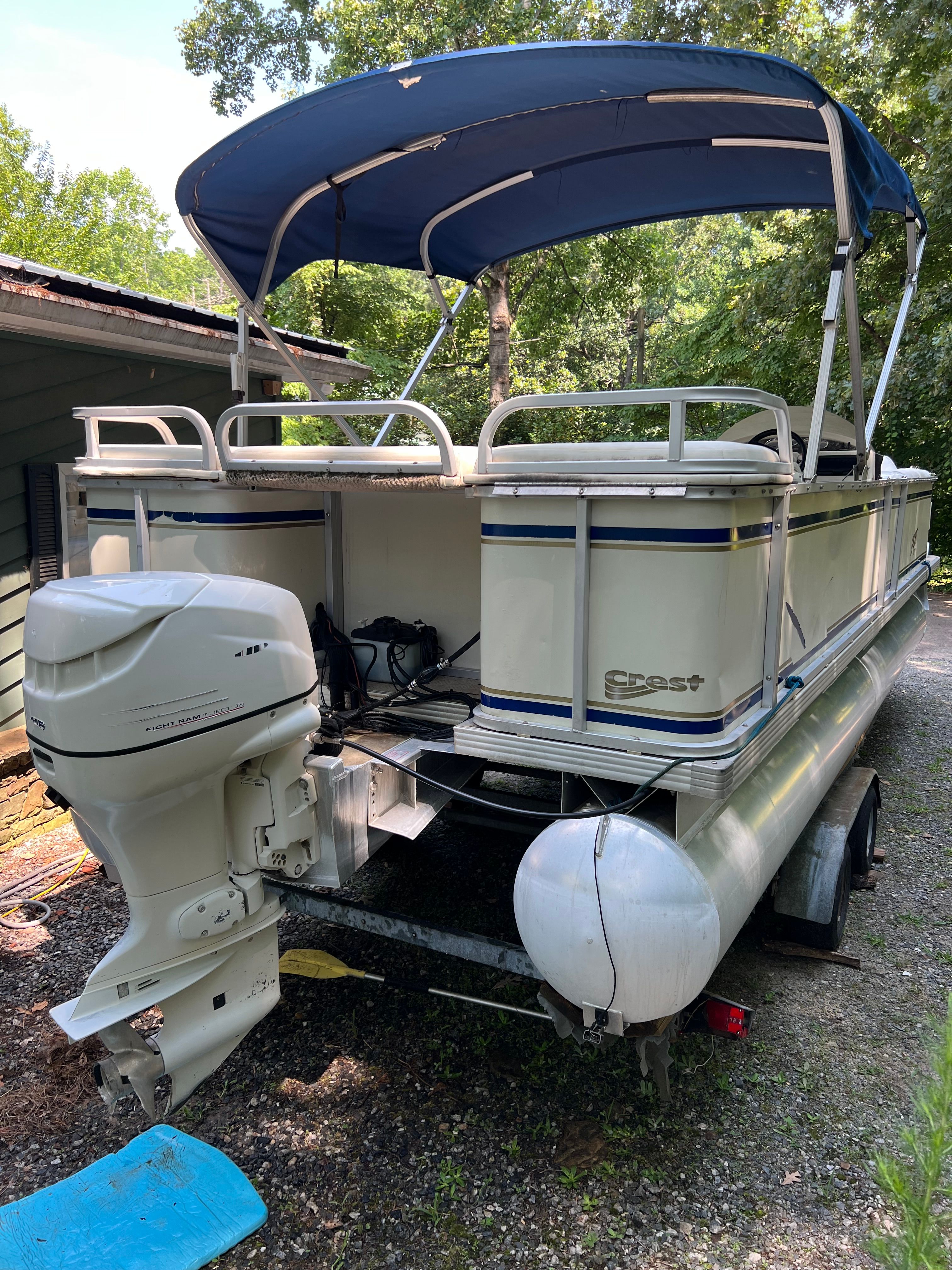 A 2003 Crest 24’ Sport pontoon boat with a blue canopy, parked on a trailer.
