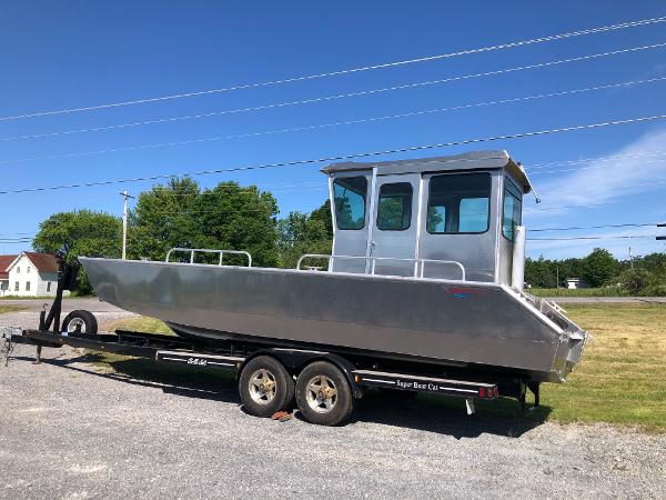 A silver workboat on a trailer, featuring a cabin and railings, parked on a gravel lot.