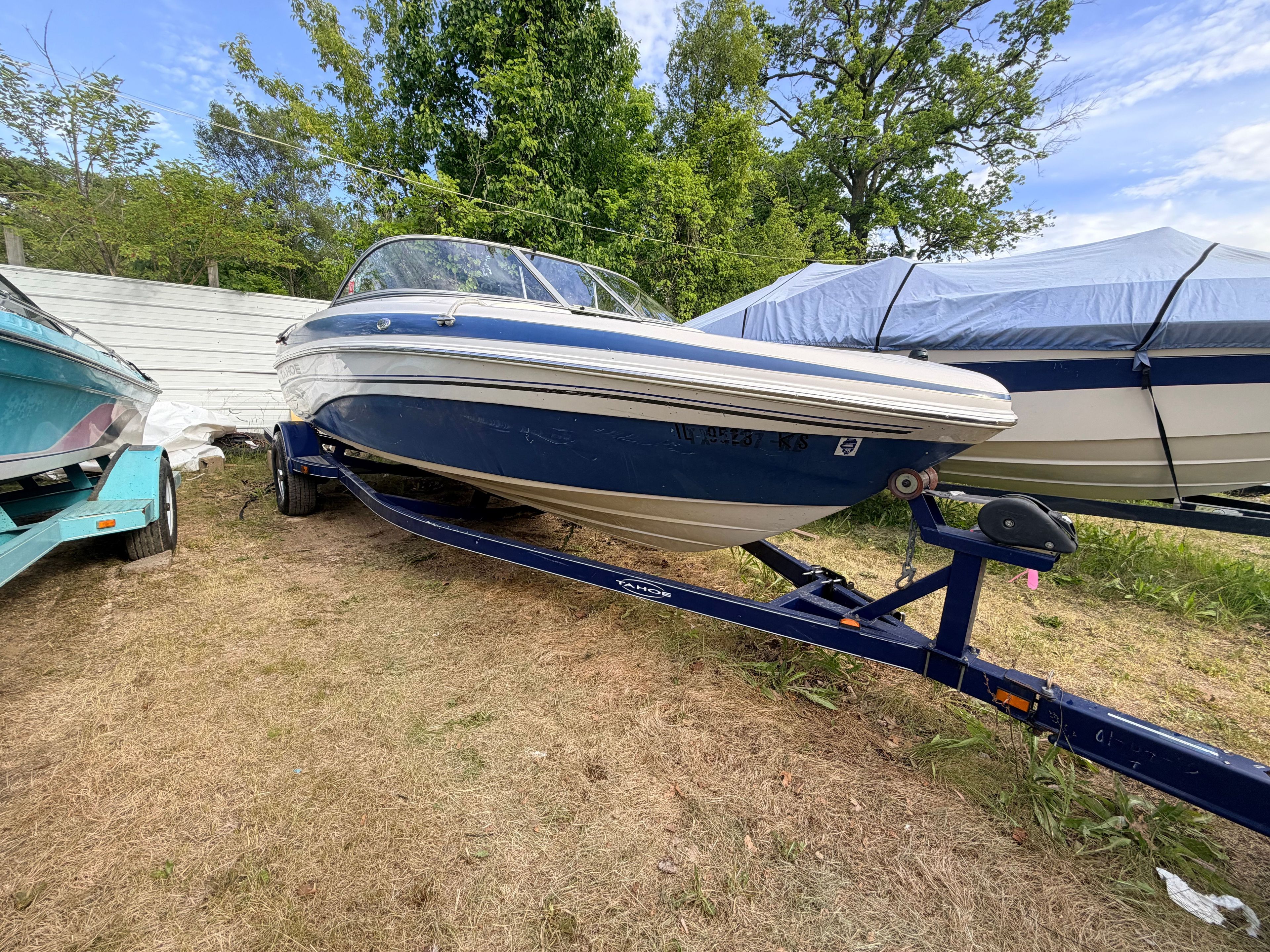 A blue and white motorboat on a trailer, surrounded by trees and other boats.