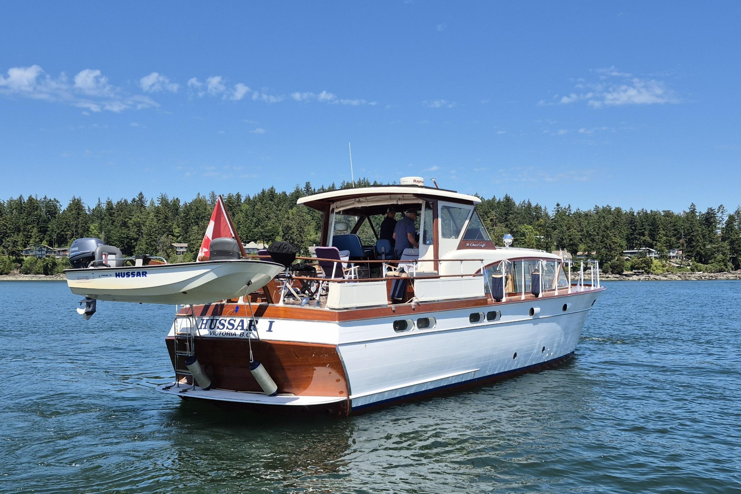 1958 Chris-Craft 500 Constellation yacht on water, with small boat attached, scenic forest backdrop.