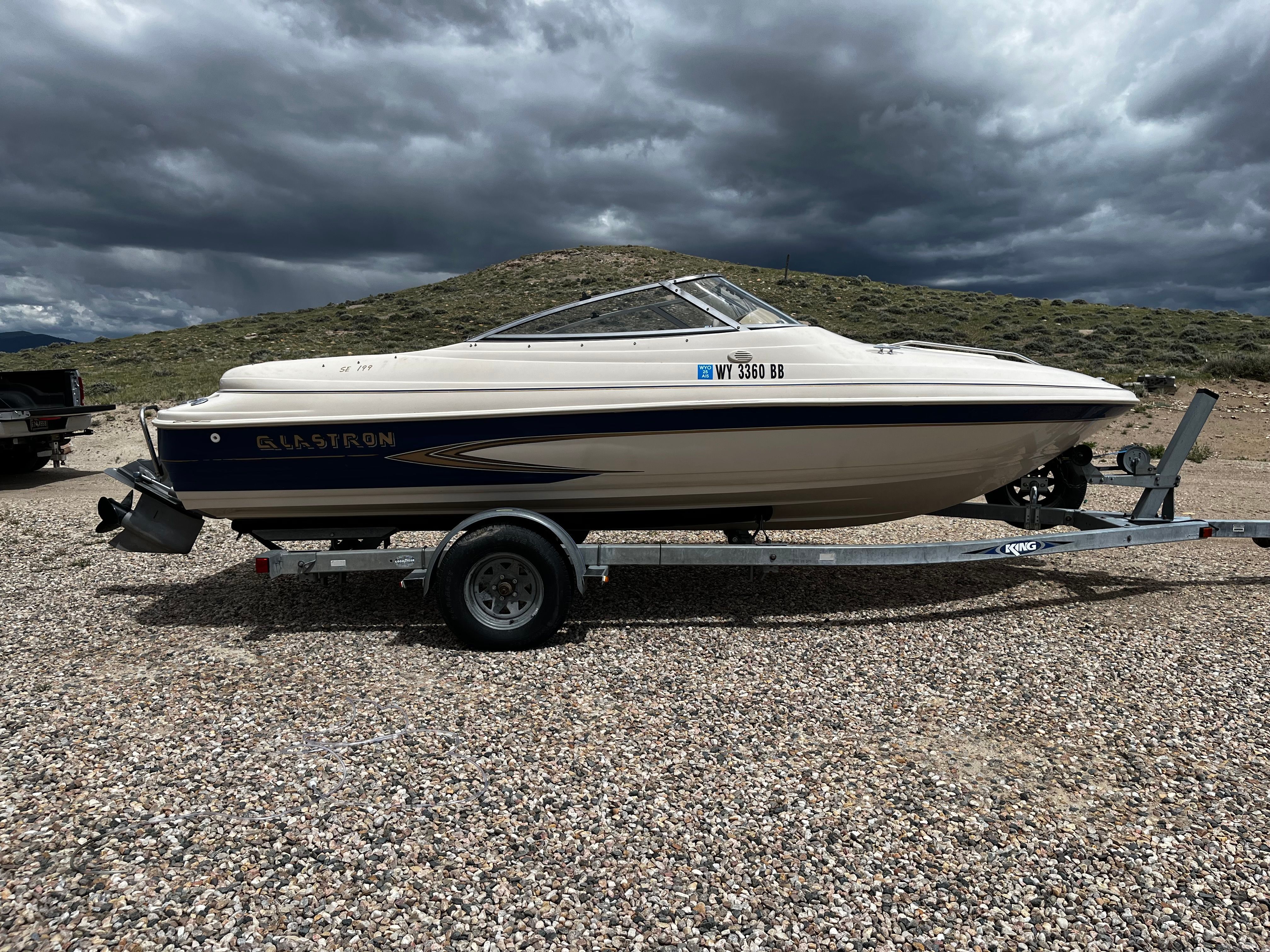 A 1998 Glastron SE199 boat on a trailer in a gravel lot under a cloudy sky.