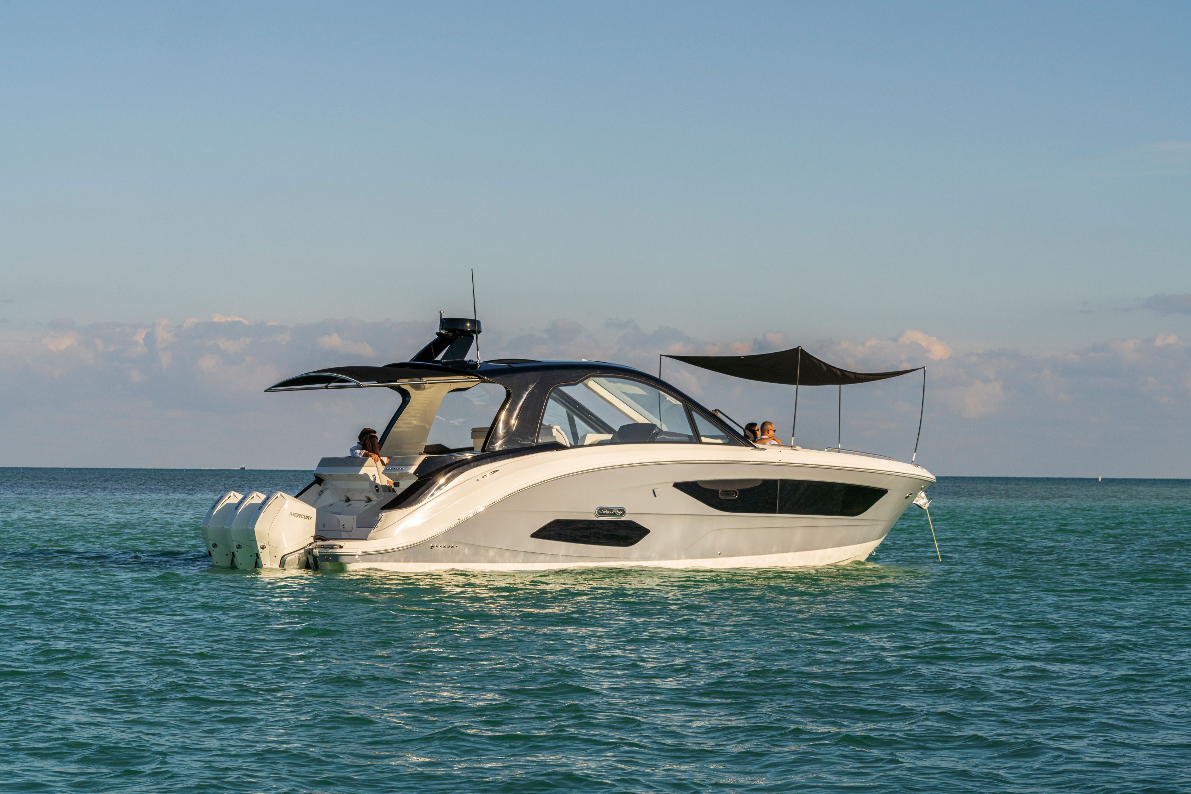 A 2026 Sea Ray Sundancer 370 Outboard on calm water with sunshades and people on board.