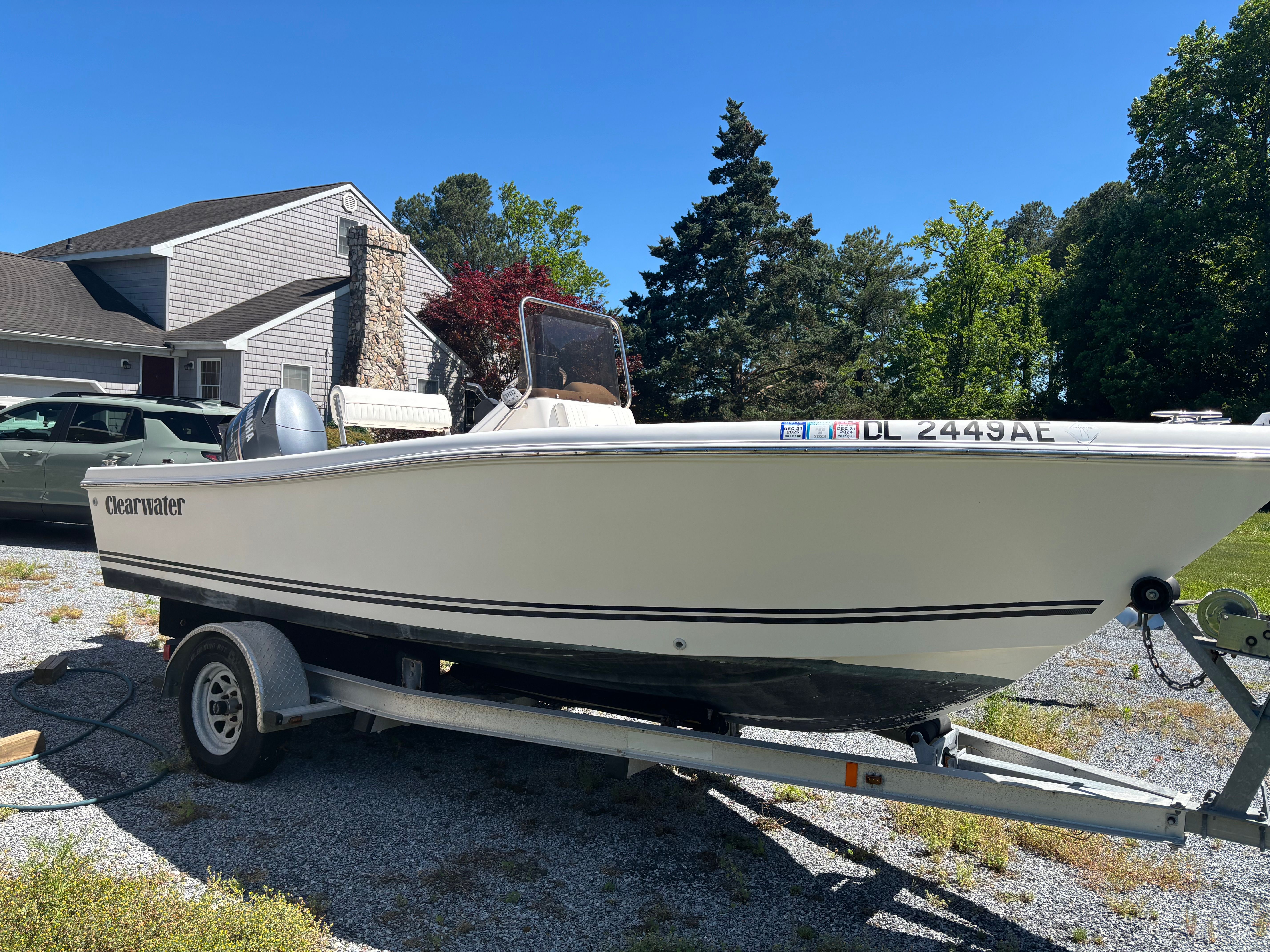 A white Clearwater boat on a trailer, parked on a gravel driveway next to a house.
