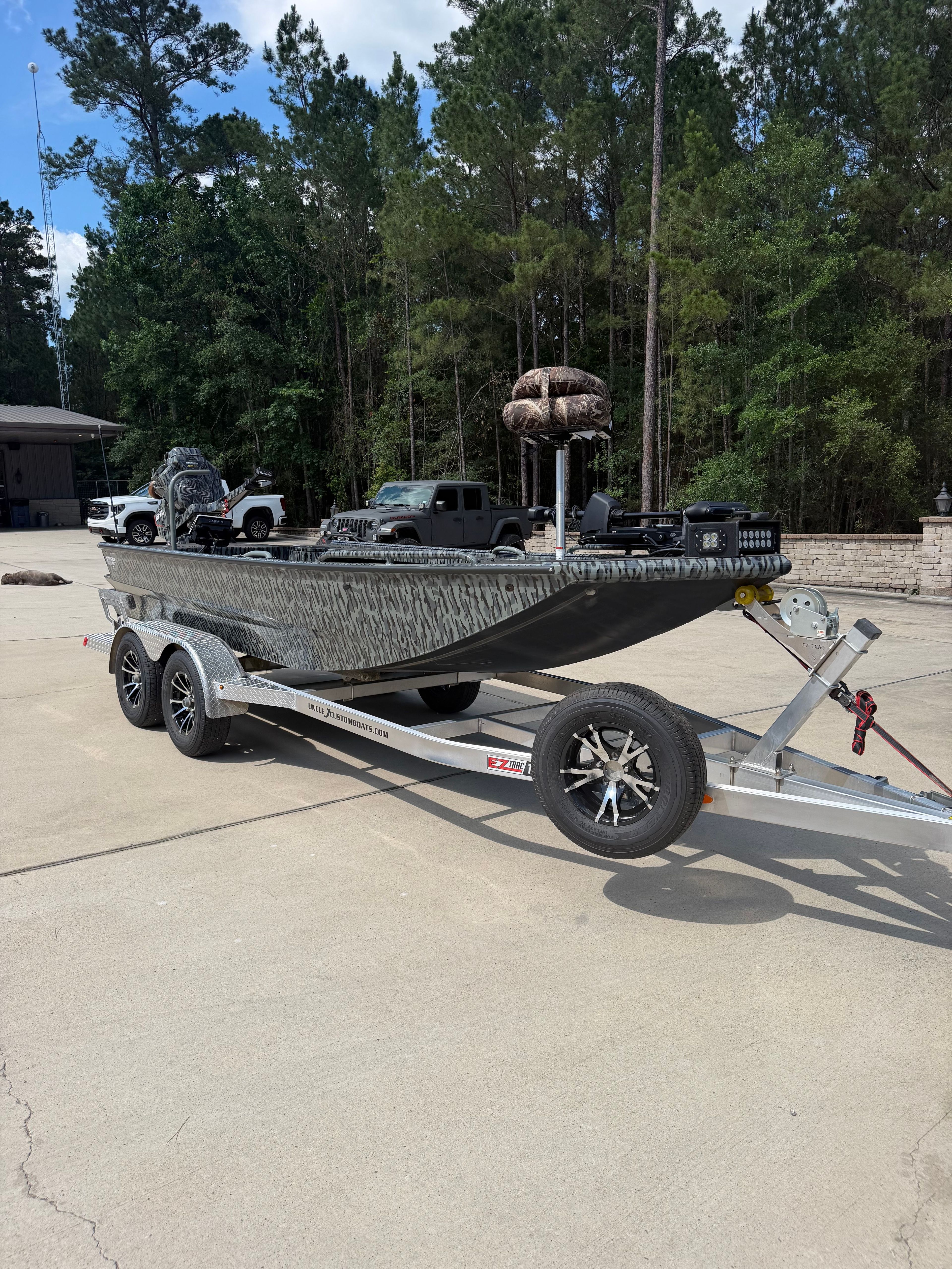 A modern boat on a trailer, surrounded by trees and vehicles in a parking area.