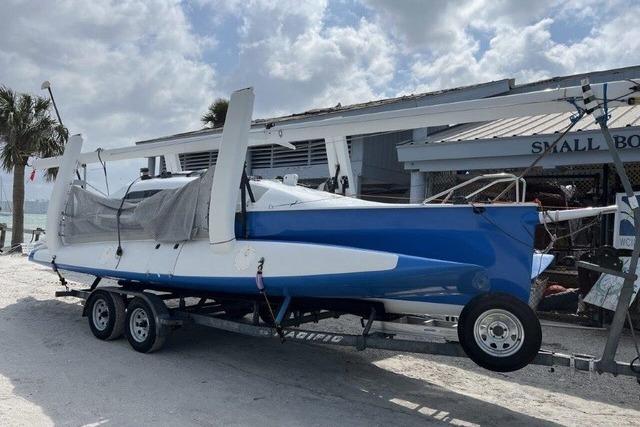 1996 Farrier F-25C trimaran on trailer near small boat shop, under cloudy sky.
