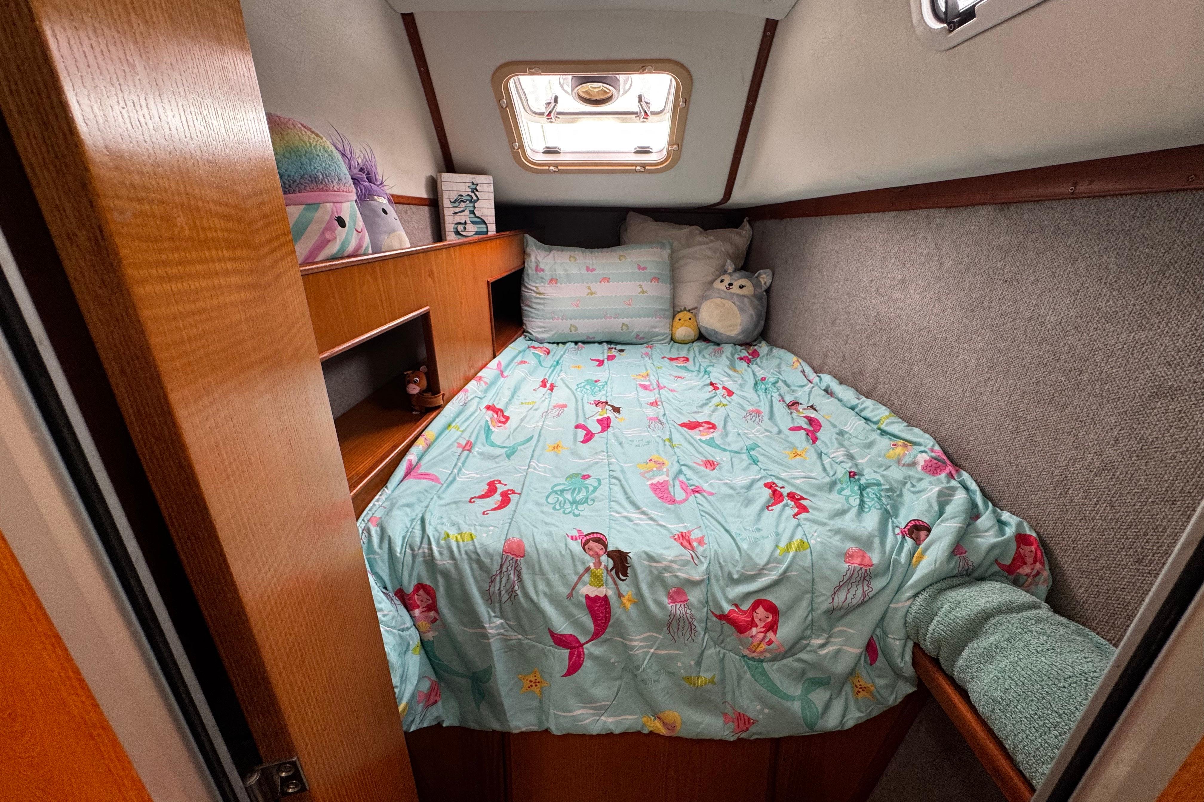 Cozy boat cabin with mermaid-themed bedding, featuring plush toys and wooden shelves.