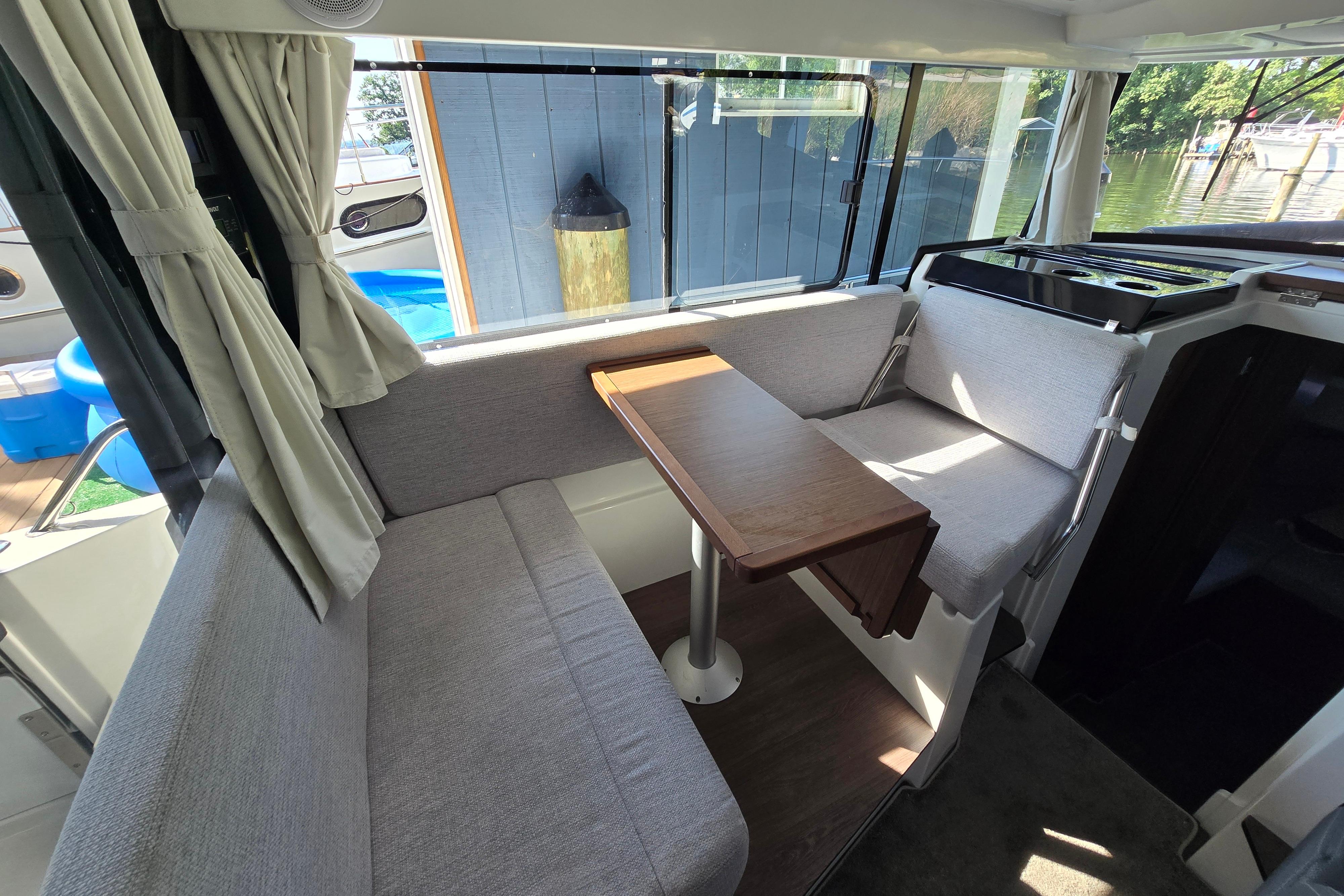Interior of a Jeanneau NC Sport 895 with gray seating, wooden table, and windows with curtains.