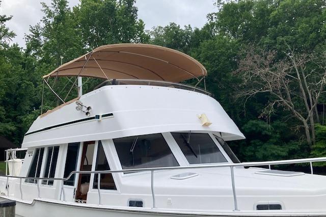 2000 Mainship 430 Trawler with canopy, docked near lush green trees.