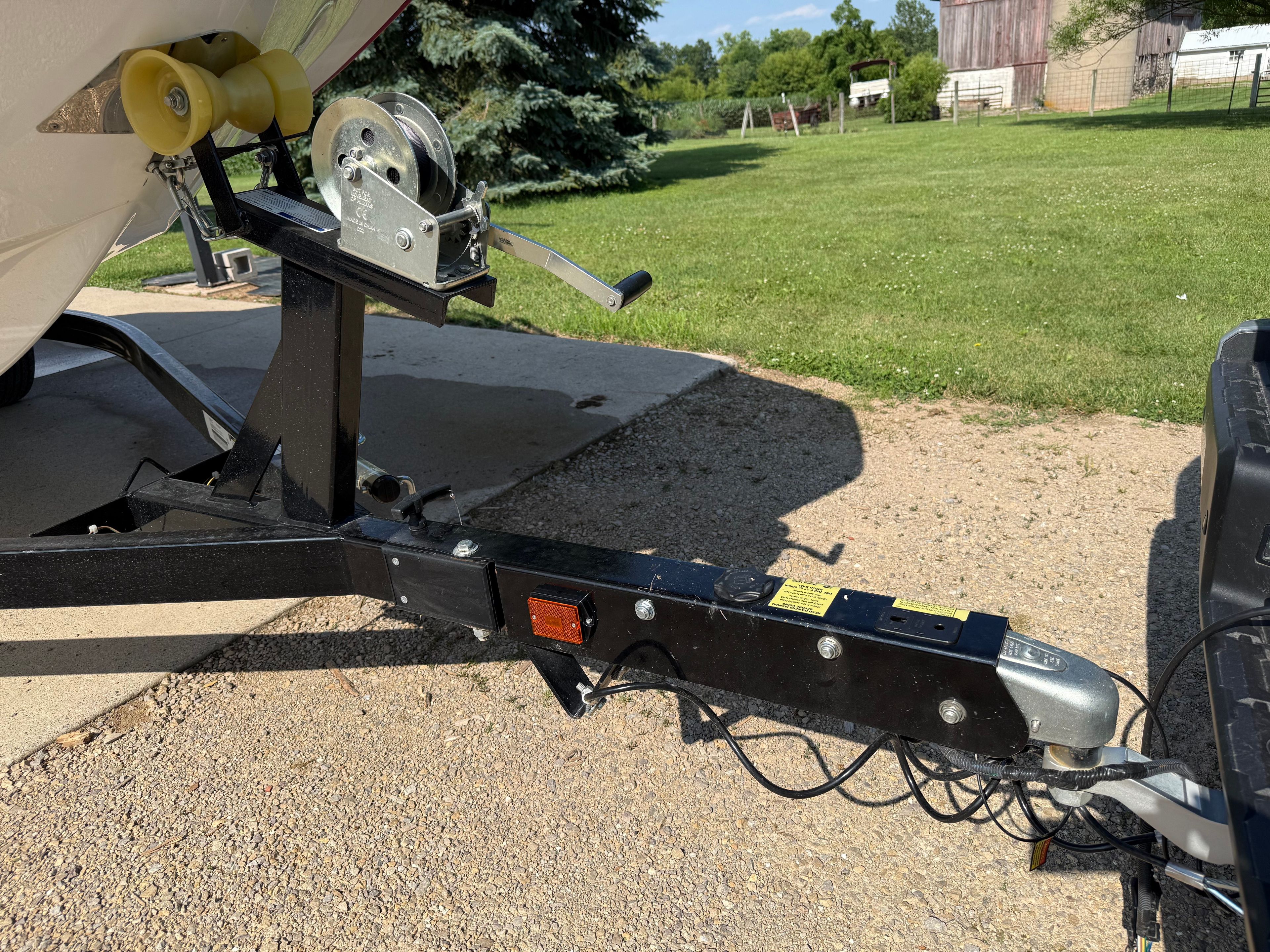 A boat trailer with a winch, parked on a gravel path next to a grassy field.